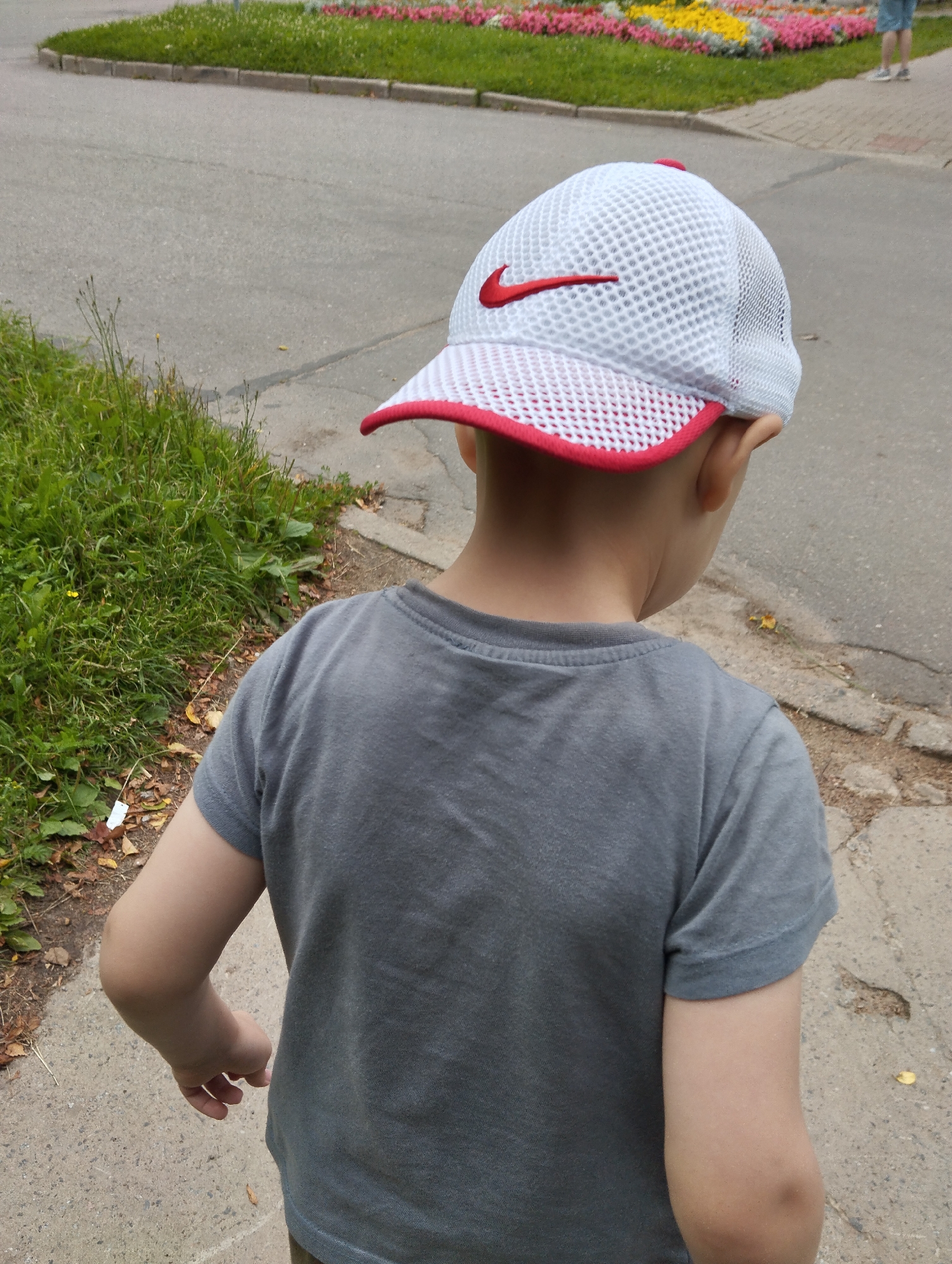 A child wearing a gray t-shirt and a white Nike cap with a red trim and logo is shown from the back. The child is walking on a paved path next to a grassy area. In the background, there are flower beds with colorful flowers and more paved areas. The scene appears to be outdoors during the daytime. The weather seems to be pleasant, with green grass and flowers in bloom. The location is Kingisepp, Russia. There is no discernible text in the image other than the Nike logo on the cap.