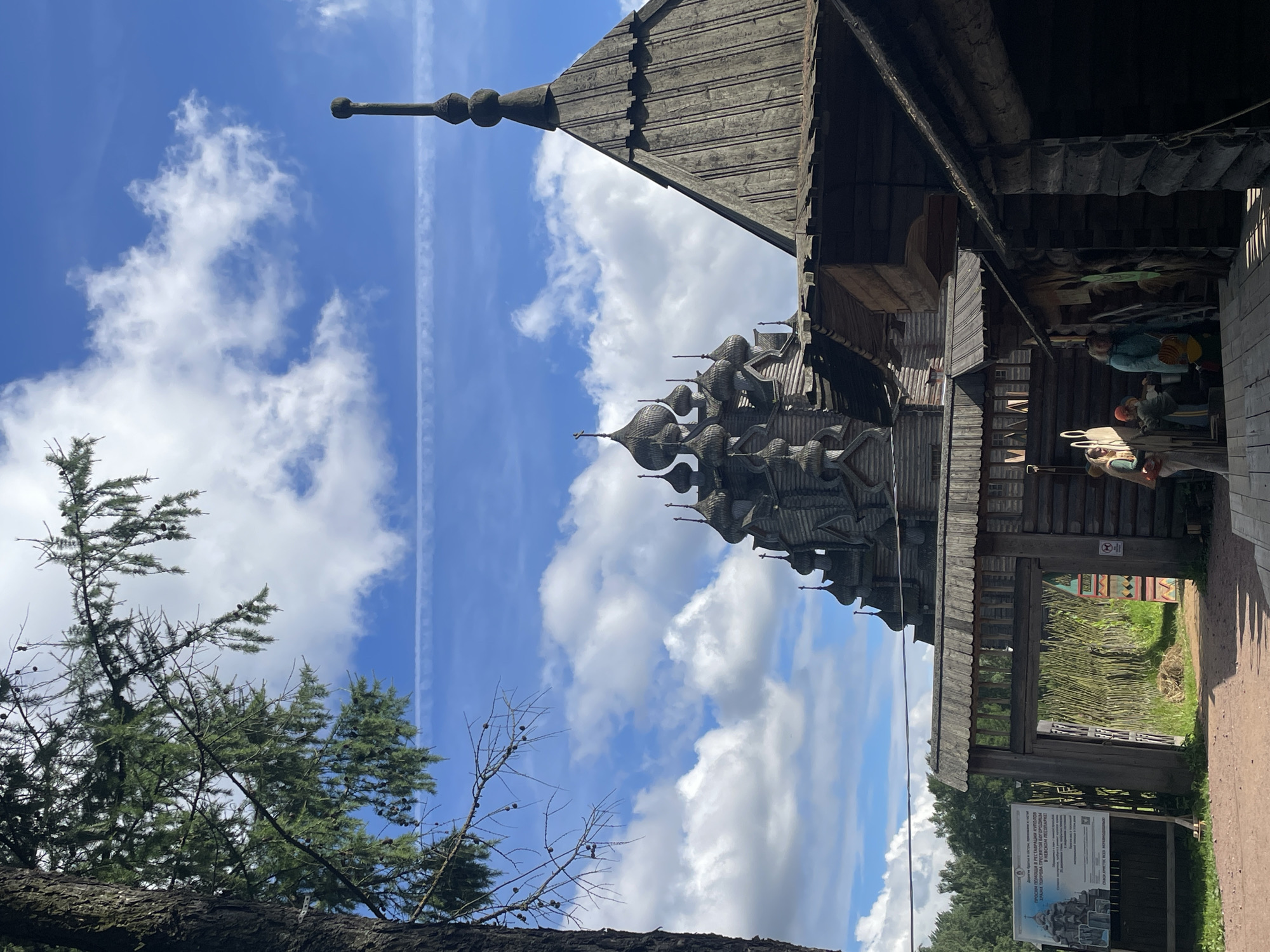 The image shows the Kizhi Pogost, a historic church complex located on Kizhi Island in Karelia, Russia, approximately 250 kilometers from Nevskii Parkleskhoz. In the foreground, a large, old tree with sparse branches partially obscures the view of the sky and the church. The sky is a bright blue with scattered white, puffy clouds and a faint contrail.
Dominating the middle ground is the Kizhi Pogost, a UNESCO World Heritage site. The most prominent structure is the Church of the Transfiguration, a massive wooden church with numerous domes and intricate wooden architecture. To the right of it, part of another wooden structure, possibly another church or a bell tower, is visible, featuring a steeply pitched roof.
In the immediate foreground, there's a rustic wooden entrance gate, and beyond it, a pathway leads towards the church complex. A signpost with Cyrillic text and an image of the Kizhi Pogost is positioned to the left of the gate. A small group of people are visible near the entrance, some standing and others appearing to be interacting with each other. One person is wearing a light-colored headscarf, and another is holding a bag. The architecture is predominantly made of dark, weathered wood. The environment suggests an outdoor historical or cultural site.