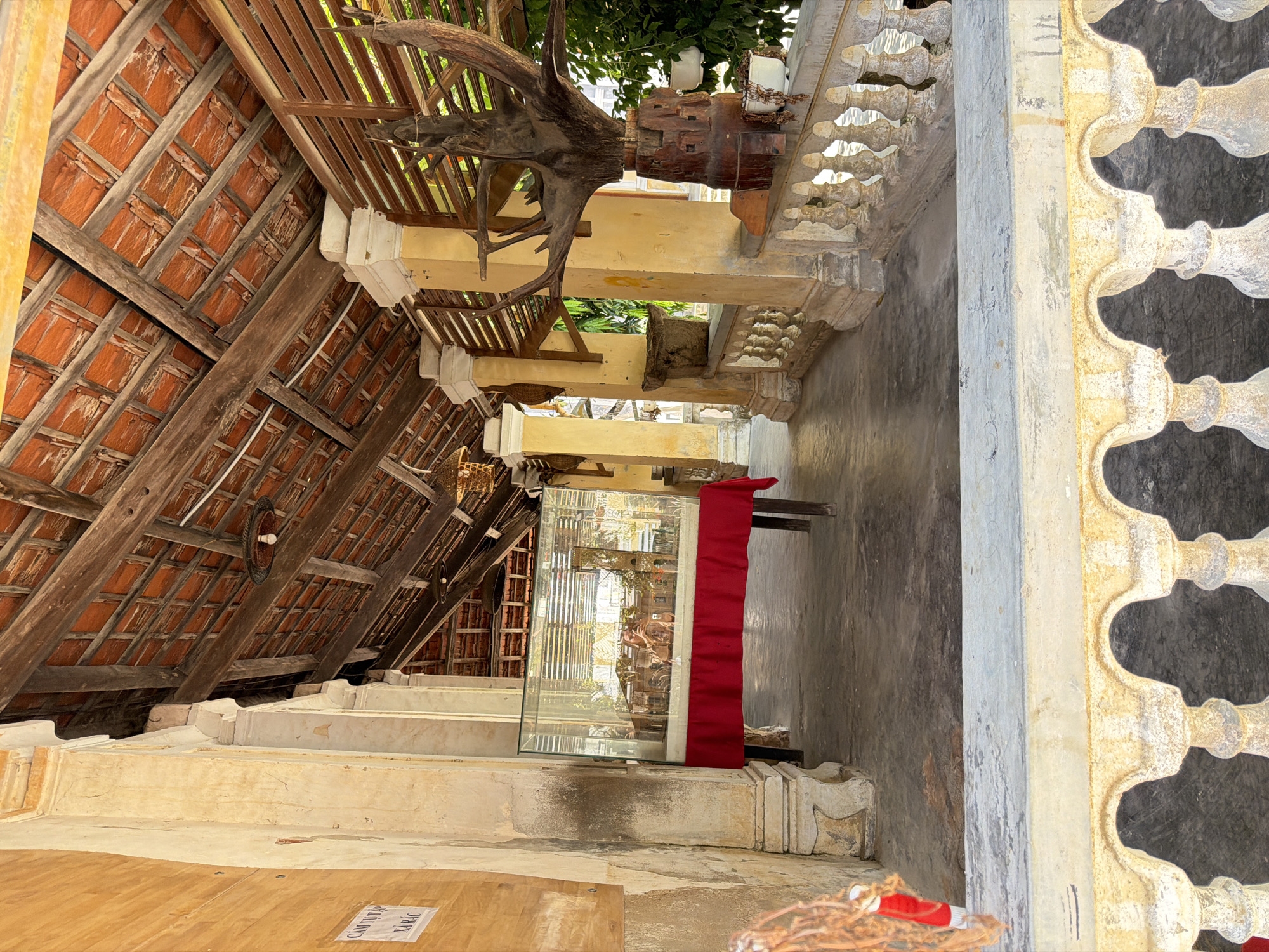 The image shows an outdoor walkway with a covered roof supported by pillars. The roof is made of terracotta tiles and wooden beams, showing signs of age. The walkway has a decorative railing with balusters. On the left, there is a display case with a model inside, covered by a red cloth on a table. Further down the walkway, there is a large, gnarled piece of wood mounted on a wooden base, with hanging planters. The walls are painted yellow and show wear. The ground is paved. A sign with Vietnamese text is attached to the left wall. The scene appears to be in Hội Tín, Vietnam, possibly a historical site or temple. There are no people visible in the image.