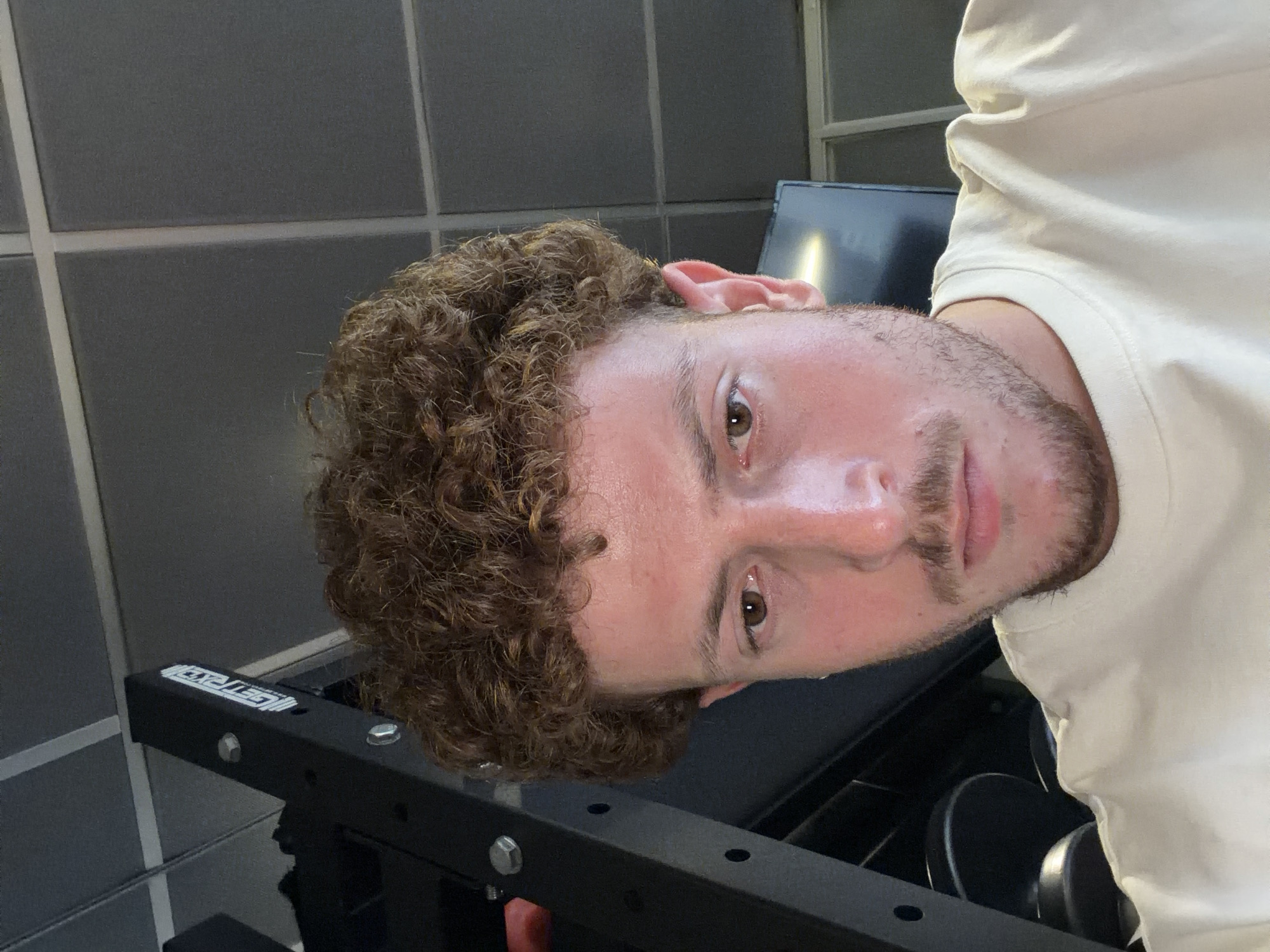 A selfie of a person with curly hair and a beard, slightly sweaty, in what appears to be a gym setting. The background includes tiled walls and gym equipment. The location is not identifiable as New York City, USA from the provided image.