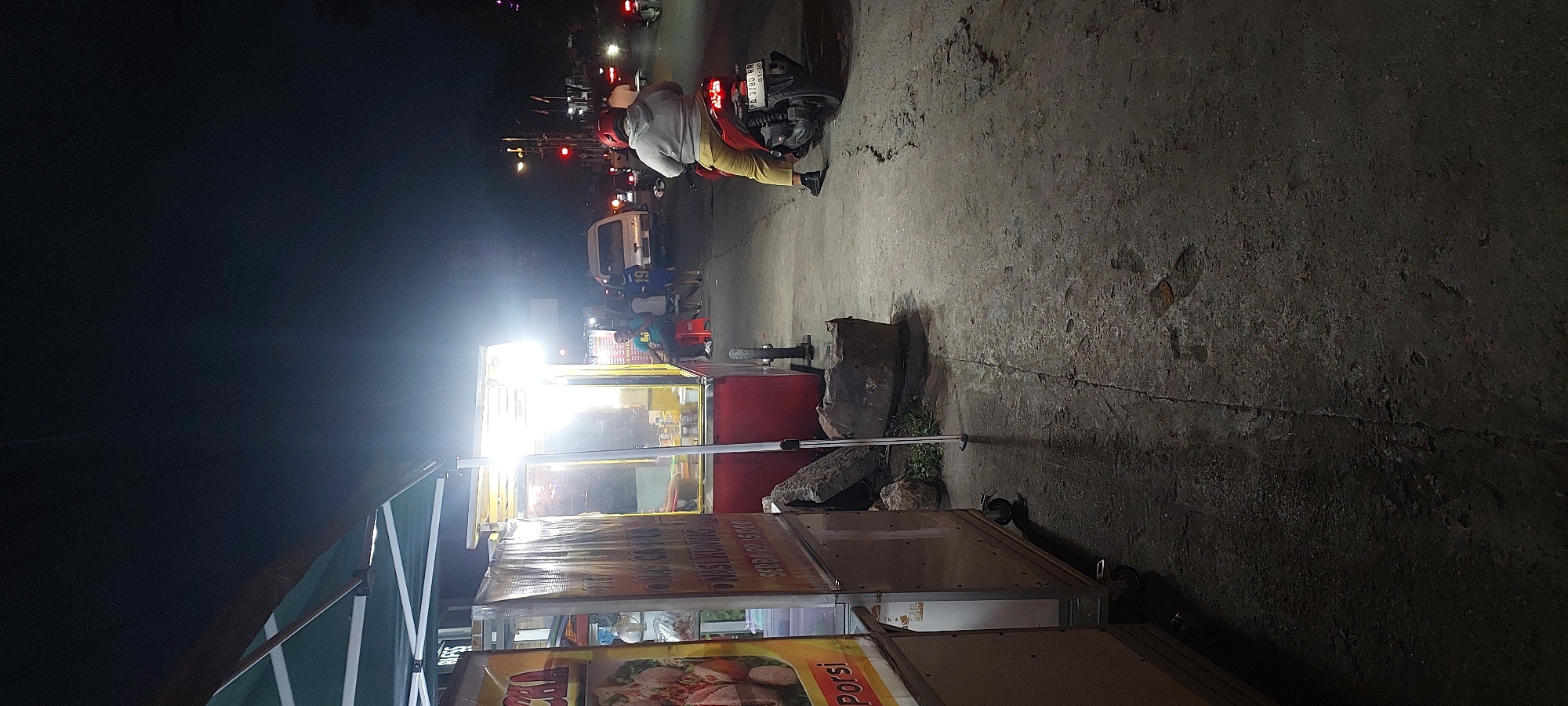 The image depicts a nighttime street scene in Jayapura, Indonesia, characterized by dimly lit food stalls and passing traffic. A red and white food cart, illuminated by a bright overhead light, is positioned on the left side of the frame. Signage on the cart, partially obscured, includes text in Indonesian. To the right of the cart, a person wearing a red helmet and yellow pants is seated on a red scooter, facing away from the camera. The scooter's license plate reads "PA 3780 RR 01-30."
The street itself is paved with concrete, showing signs of wear and tear, with visible cracks. Several large rocks are placed near the food cart. In the background, other vehicles, including a white van and several motorbikes, are visible on the road, with their headlights and taillights creating streaks of light. The overall atmosphere is that of a typical urban night market or roadside eatery. The dark sky suggests it is late at night.