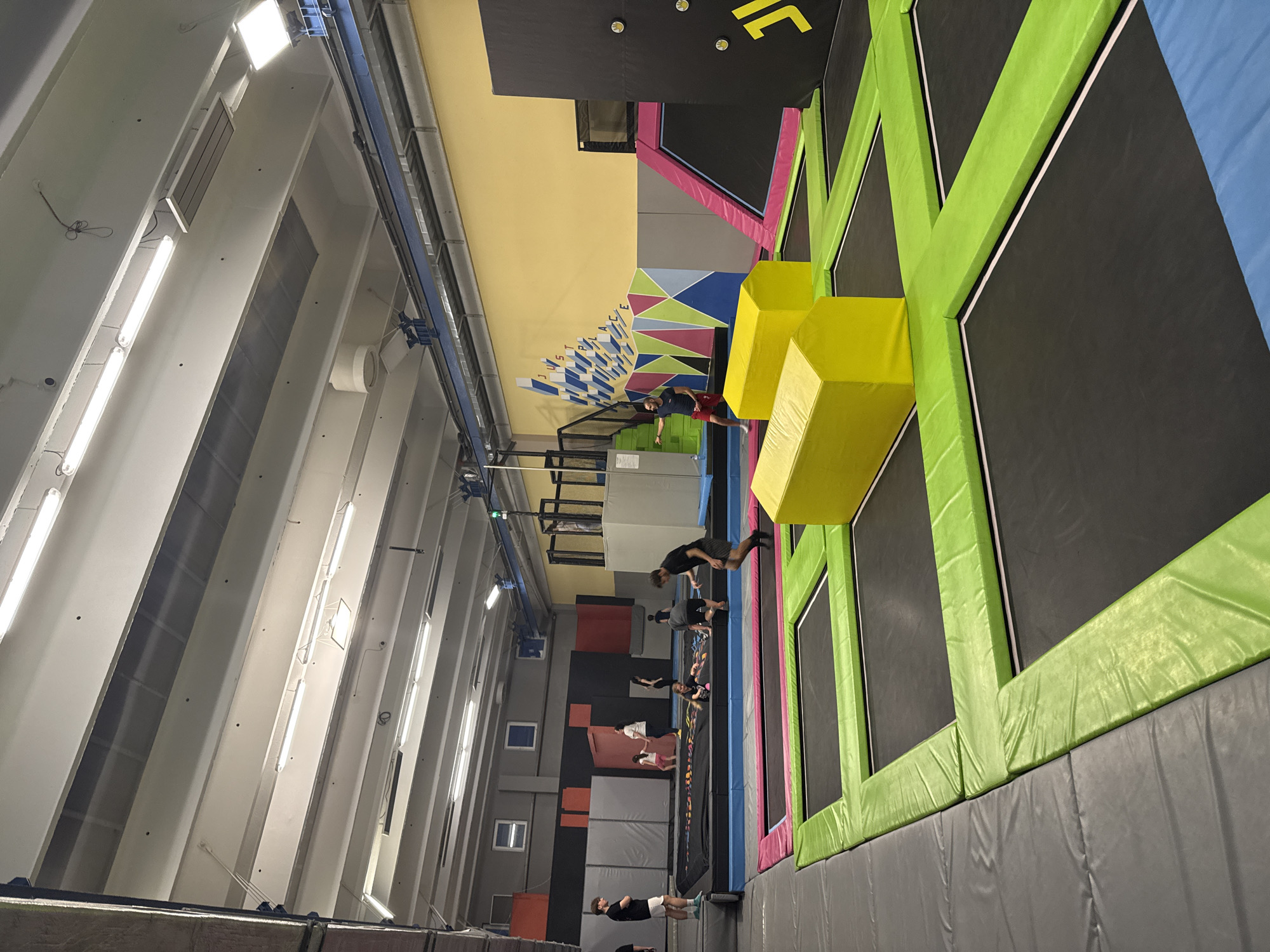 A photograph depicts the interior of a trampoline park, potentially located in Sofia, Bulgaria. The scene is filled with interconnected trampolines featuring black jumping surfaces and safety pads in green, blue, and pink. A structure of stacked yellow foam blocks is positioned adjacent to one of the trampolines.
Several people are present within the trampoline park. One individual is in mid-air, seemingly jumping towards the yellow blocks, while others are either standing or jumping on the trampolines.
The building's interior features a high ceiling with visible overhead lighting fixtures and support beams. The walls are decorated with a variety of colorful geometric patterns. Gray padded walls line the bottom of the trampolines.