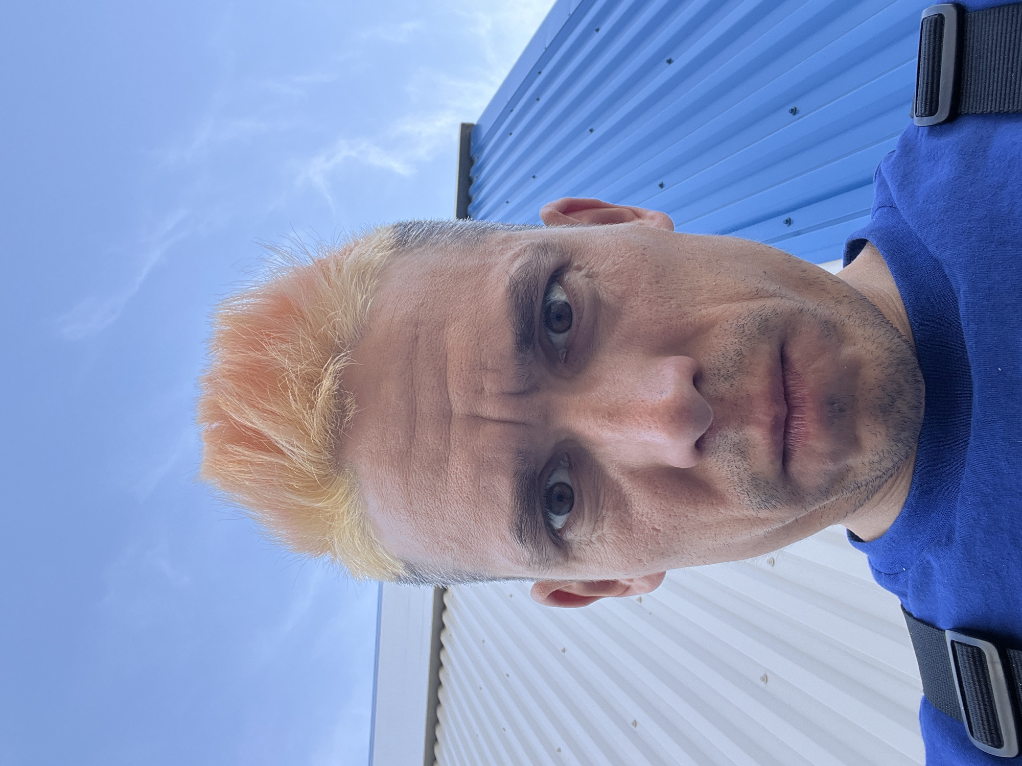 A close-up, low-angle shot of a Caucasian man with short, light-colored hair, standing outdoors. His hair appears to be bleached with some orange-ish highlights. He has gray eyes and stubble. He wears a blue T-shirt and a black strap or belt is visible on his shoulder.
Behind him, are the corrugated metal walls of buildings. One section is painted blue, another section is painted white. The sky is visible above the buildings, with a few light clouds. The location of the image is unknown and can't be determined from the given image. There is no specific information in the image that verifies it is Chaikovsky, Russia.