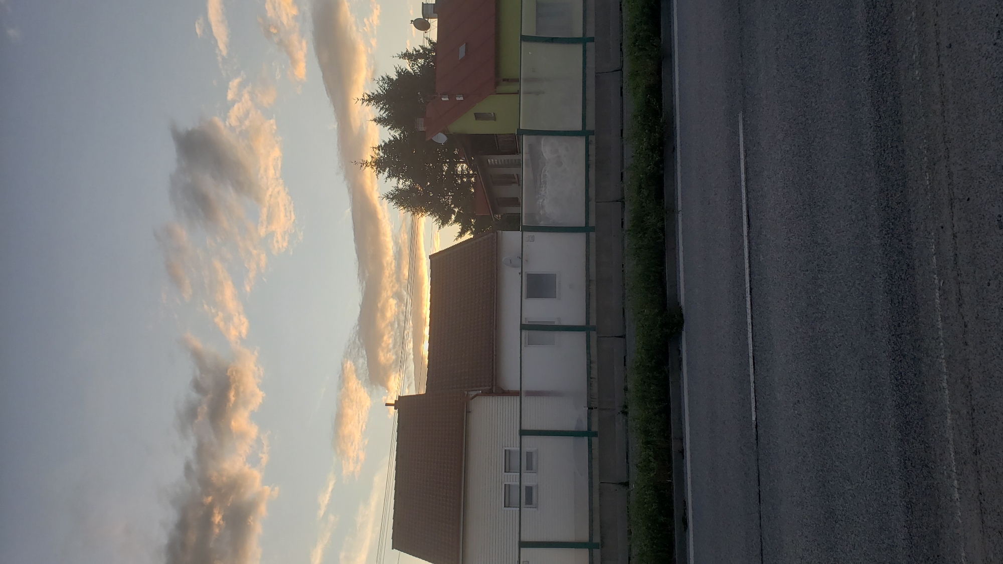 The image depicts a suburban scene with residential buildings and a highway in Prešov, Slovakia, under a cloudy sky during what appears to be sunset or sunrise. In the foreground, a dark asphalt road with a white lane marker is visible. Behind the road, a strip of green grass separates it from a low, transparent barrier with green framing. Beyond this barrier are several houses with red and green tiled roofs. The buildings are of various sizes and styles, with some featuring white facades and others with darker exteriors. Trees, including tall evergreens, are situated behind the houses. The sky is filled with a mix of light blue and scattered clouds, some of which are illuminated with warm tones, suggesting the sun is low on the horizon. There are no people visible in the image. The overall impression is of a quiet residential area adjacent to a transportation route, under a dramatic sky.