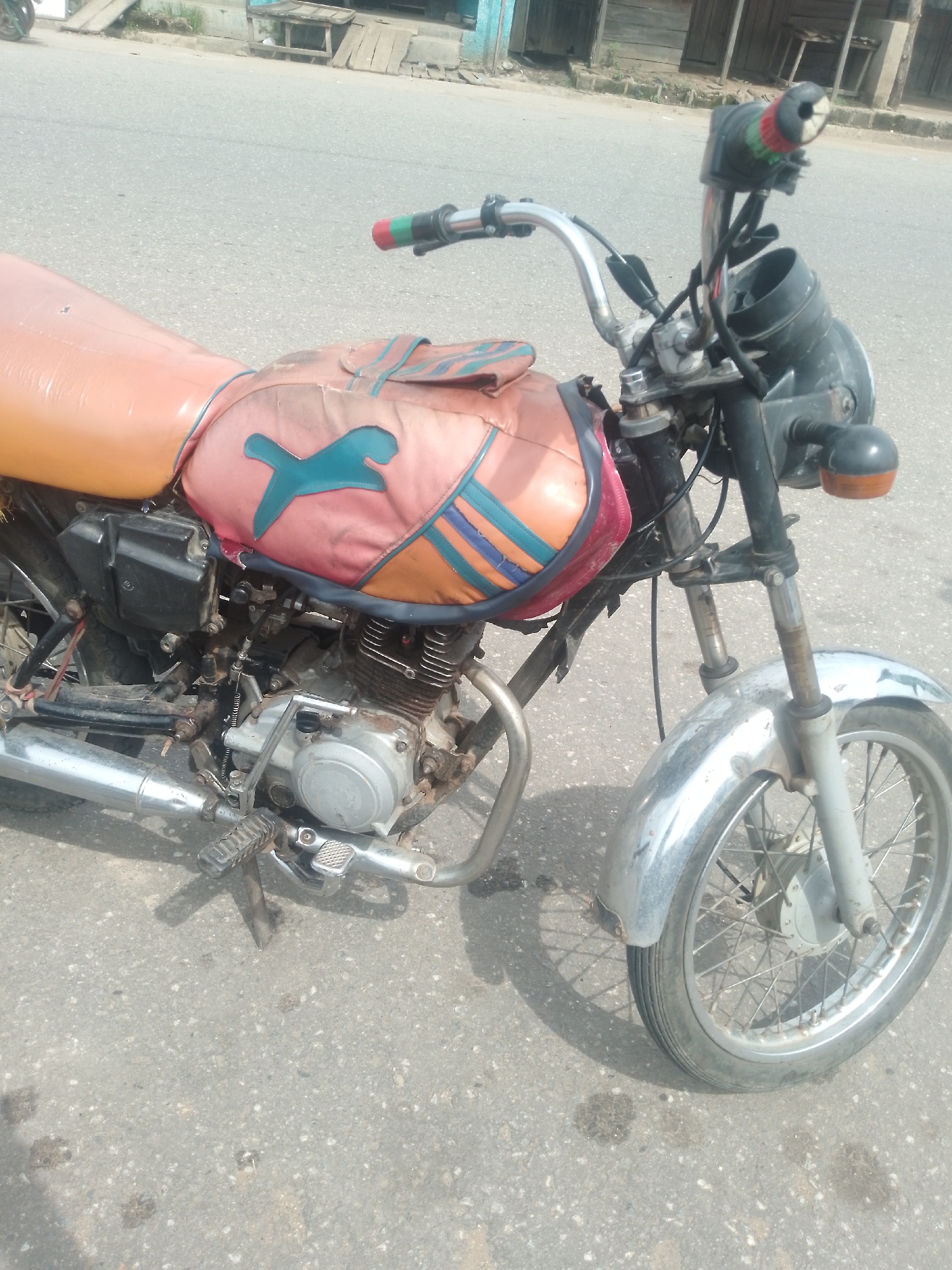 In the city of Serti, Nigeria, a motorcycle with an orange seat and a pink and teal fuel tank cover is parked on an asphalt road. The motorcycle has a silver exhaust pipe, a chrome front fender, and a black headlight. The motorcycle's engine is visible, and it appears to be a manual transmission. The road is cracked and has some oil stains. In the background, there are wooden structures that appear to be shops or stalls. The lighting suggests it is daytime, with shadows cast on the road.