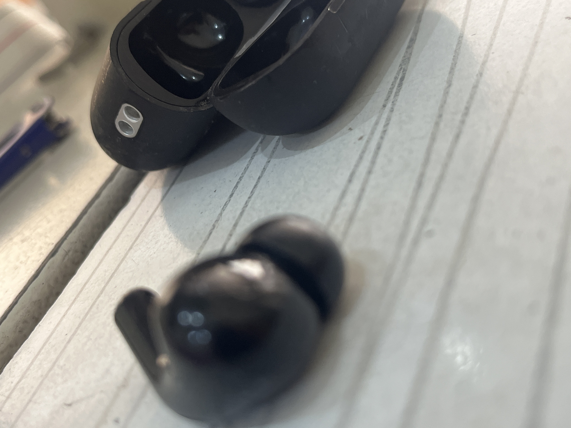 The image shows a close-up view of black wireless earbuds and their charging case, placed on a surface with parallel lines, possibly a desk or a notebook. One earbud is detached from the case and positioned on the left side of the frame, appearing slightly blurred. The charging case is open and positioned to the right, revealing the interior where earbuds are typically stored. The case has two small, silver oval indicators on its exterior. The lighting in the image creates reflections on the glossy surfaces of the earbuds and case, and casts shadows on the lined surface. The background is out of focus, with some blurry shapes that might indicate other objects or a wall. There are no people or animals visible in the image.