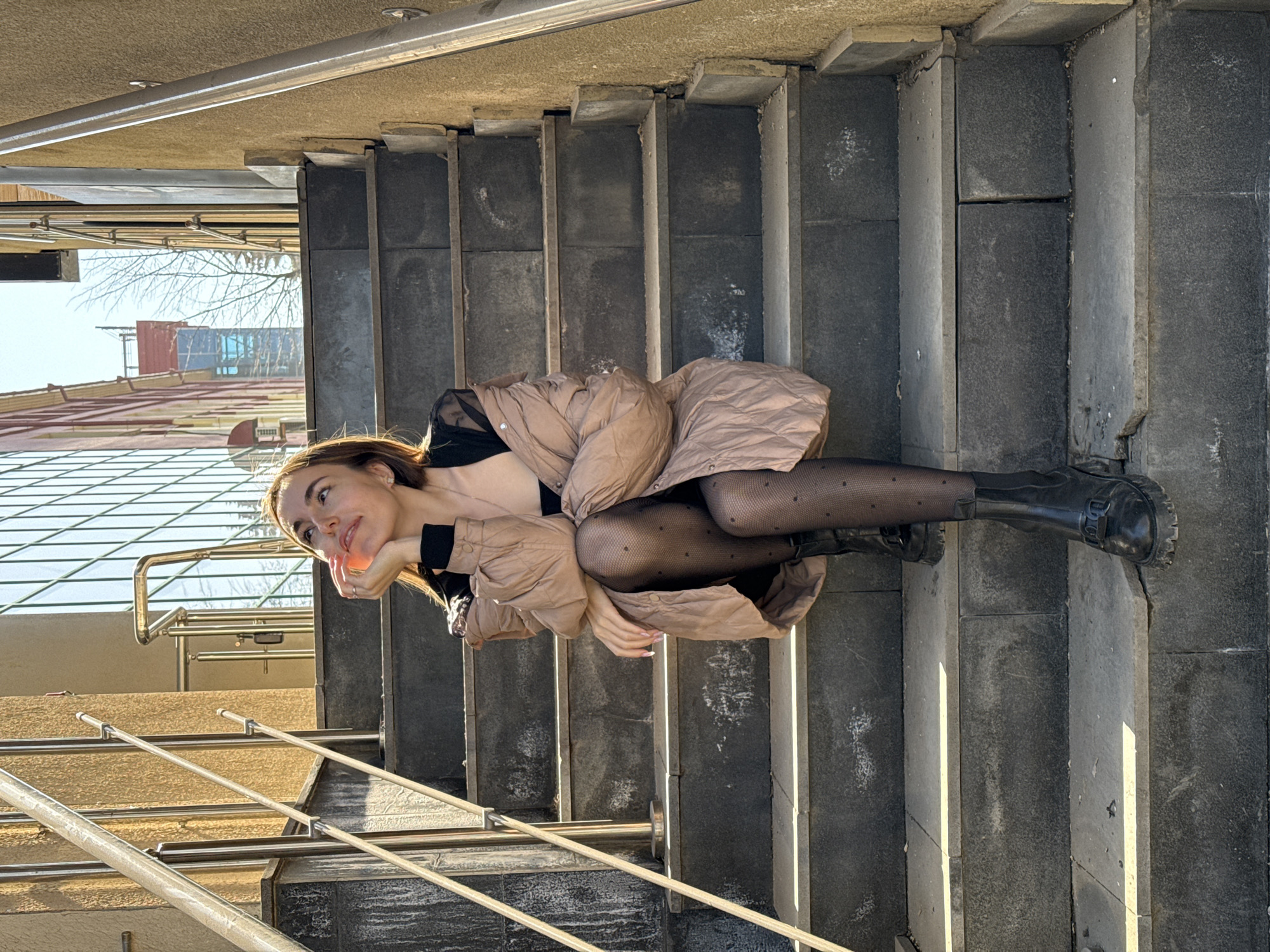 A young woman with long brown hair is lying on a set of concrete stairs. She is wearing a black top, patterned tights, black boots, and a beige puffer jacket. Her head is resting on her hand, and she is looking to the left. A metal railing is visible on the left side of the frame. A building and bare trees are in the background. The location is Izhevsk, Russia.