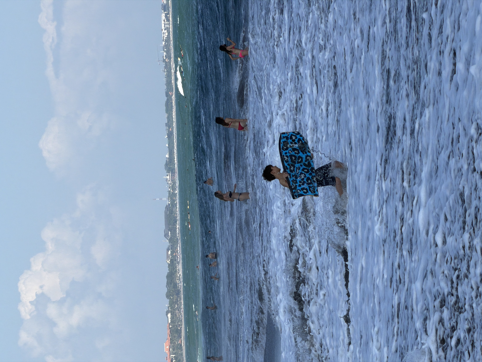 In North Kuta, Indonesia, on a wavy beach, a boy in black shorts runs through foamy white water, carrying a blue and black leopard-print bodyboard. Several people are in the ocean, including a woman in a black bikini and a woman in a red bikini. In the distance, a line of buildings and palm trees borders the shoreline under a cloudy blue sky.
