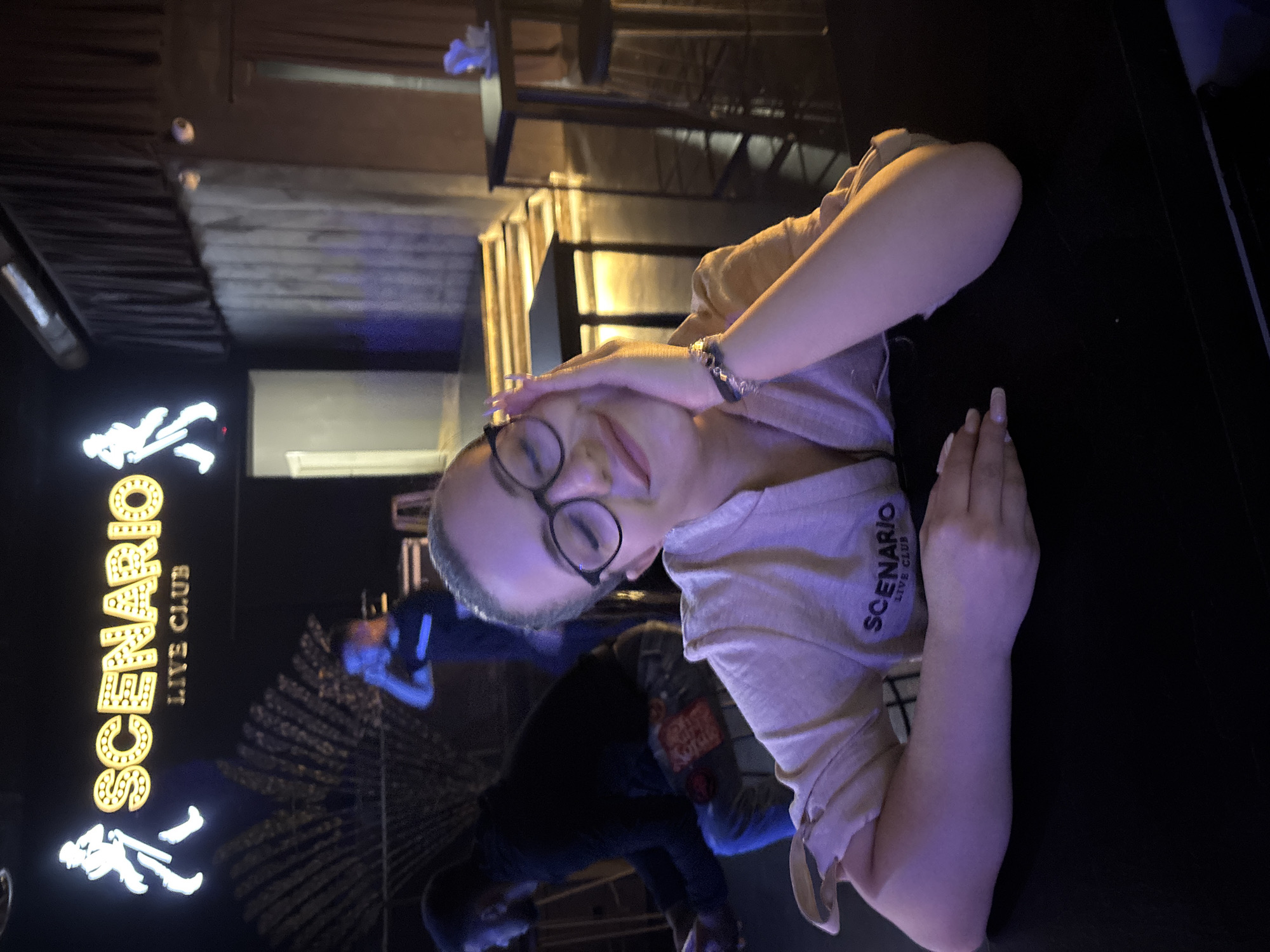 The image shows a young woman with glasses and light-colored clothing, resting her head on her hand. She is wearing a light pink garment with the text "SCENARIO LIVE CLUB" visible on the left side of her chest. The background appears to be a dimly lit club or bar with a neon sign that reads "SCENARIO LIVE CLUB" with a silhouette of a walking figure to the left of the text. There are other people in the background, including one wearing a dark shirt and another in a black hoodie. The environment suggests an indoor entertainment venue, possibly a nightclub, in Tsarevo, Bulgaria. The lighting is moody, with purple and yellow hues, common in such establishments.