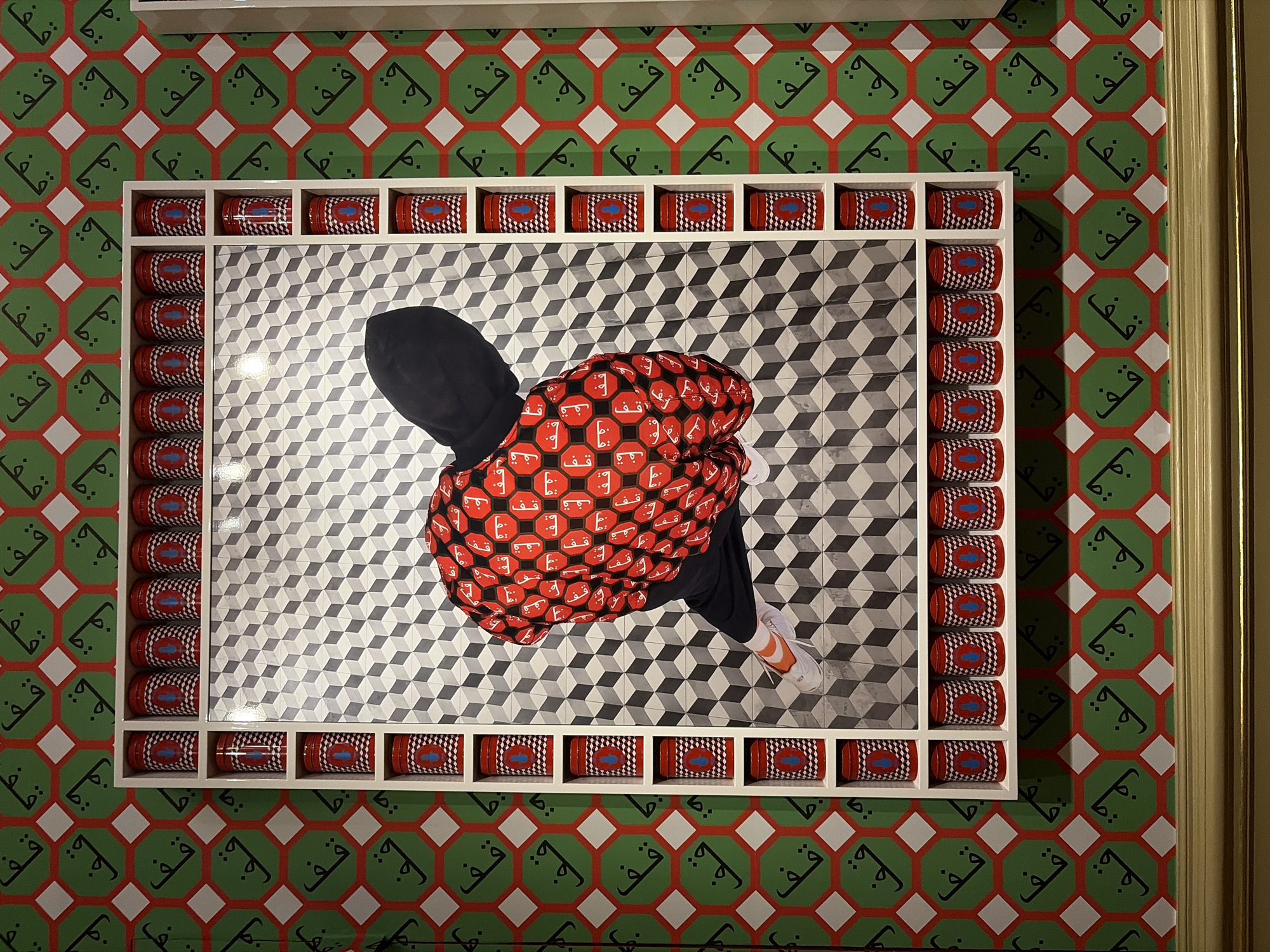 The image shows a framed artwork in a room with distinctive wallpaper. The wallpaper features a repeating pattern of green octagons outlined in red, with a black Arabic-like calligraphy symbol inside each octagon. The frame of the artwork is a white rectangular structure with multiple shelves. These shelves are filled with rows of identical red cylindrical objects, each decorated with a blue graphic of a hand with an eye in the palm.
The artwork itself is a portrait of a person from behind, viewed from a high angle. The person is wearing a black hoodie and a red jacket covered in a repeating pattern of white symbols. The background of the artwork is a tiled geometric pattern of black, white, and gray squares arranged to create an optical illusion of cubes. The person is wearing dark pants and white socks with orange accents on their shoes.
This artwork is displayed at the Mauritshuis museum in The Hague, Netherlands, as part of the "Modern Times" exhibition, which features contemporary art juxtaposed with the museum's classical collection. The specific artwork is titled "Untitled (2023)" by artist Fana.