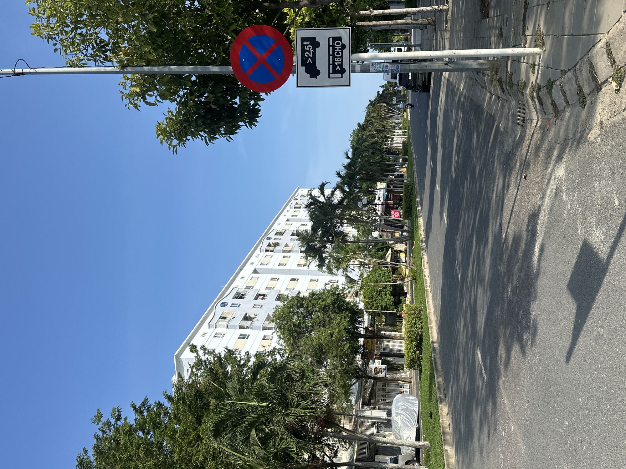 The image depicts a street scene in Nha Trang, Vietnam, likely on a clear, sunny day. The perspective is from a low angle, looking down a paved road lined with trees and buildings.
In the foreground, the asphalt road shows shadows cast by trees. A concrete sidewalk with some vegetation growing through cracks runs along the right side of the road. A utility pole stands on the sidewalk, equipped with two traffic signs. The upper sign is a red circle with a blue background and a red X, indicating "No Stopping." The lower sign is a white rectangle with black graphics. It features an outline of a truck with text "> 2,5t" above it, and a bus with text ">16 CHO" below it. These signs likely prohibit stopping for vehicles over 2.5 tons and buses with more than 16 seats.
On the left side of the road, a multi-story white building dominates the background, with visible windows and balconies. Several palm trees and other leafy trees are planted along the sidewalk in front of the building and along the street. A white car covered with a tarp is parked in front of a low-rise building that appears to house shops or businesses.
Further down the road, the street curves slightly to the right, and several other vehicles, including a dark SUV and a scooter, are visible. More trees and buildings line the street in the distance, under a clear blue sky. The overall impression is of a well-developed urban environment with a mix of residential and commercial structures, and abundant greenery.