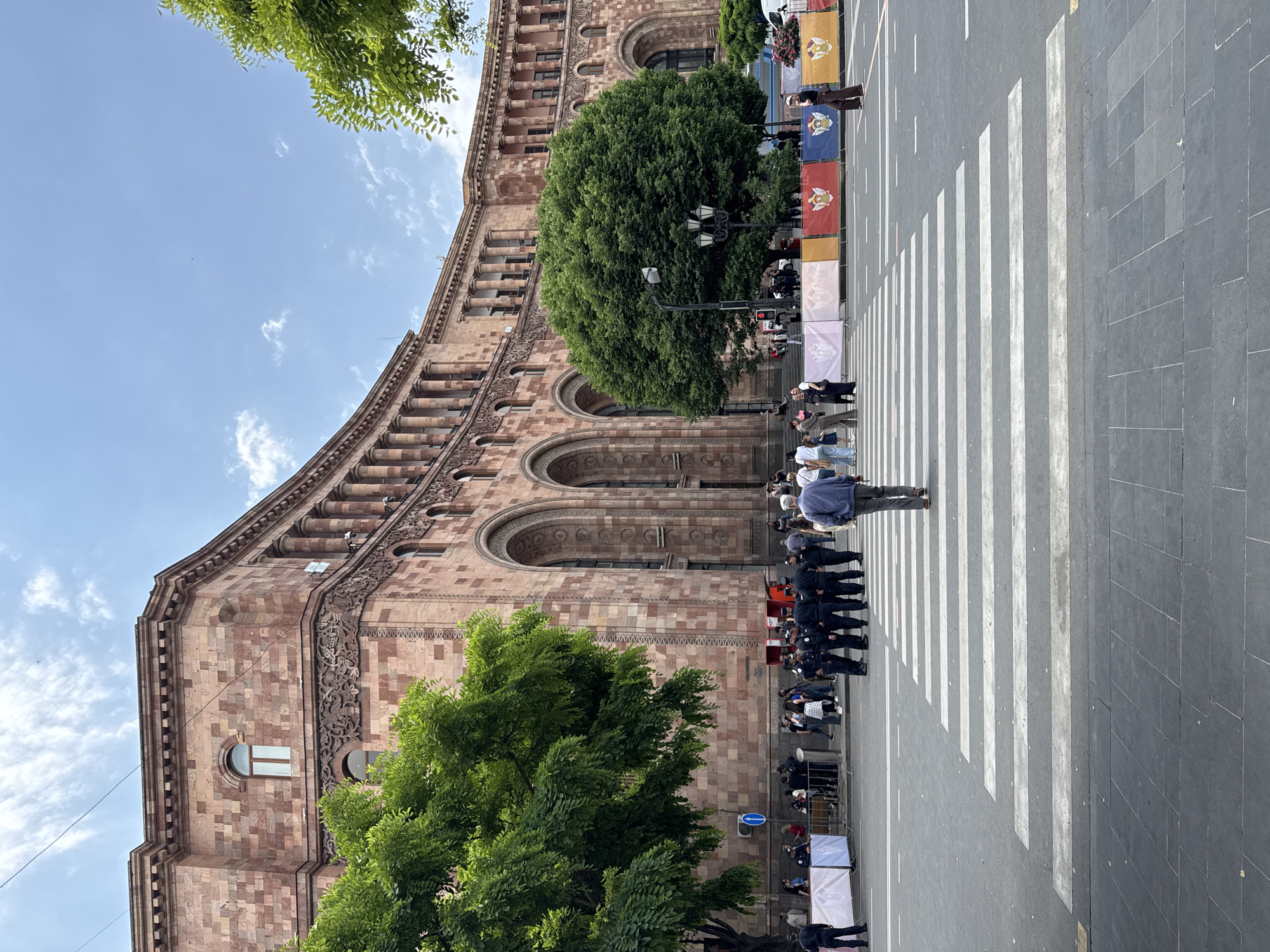 The image shows a street scene in Yerevan, Armenia, featuring a large building with ornate stonework, likely a government or historical building. A crosswalk spans the street in front of the building. People are standing along the sidewalk and in the crosswalk, some appearing to be observing an event. There are a series of flags or banners lining the street near the crosswalk. Trees are present on both sides of the building, partially obscuring its lower sections. The sky is blue with scattered clouds.