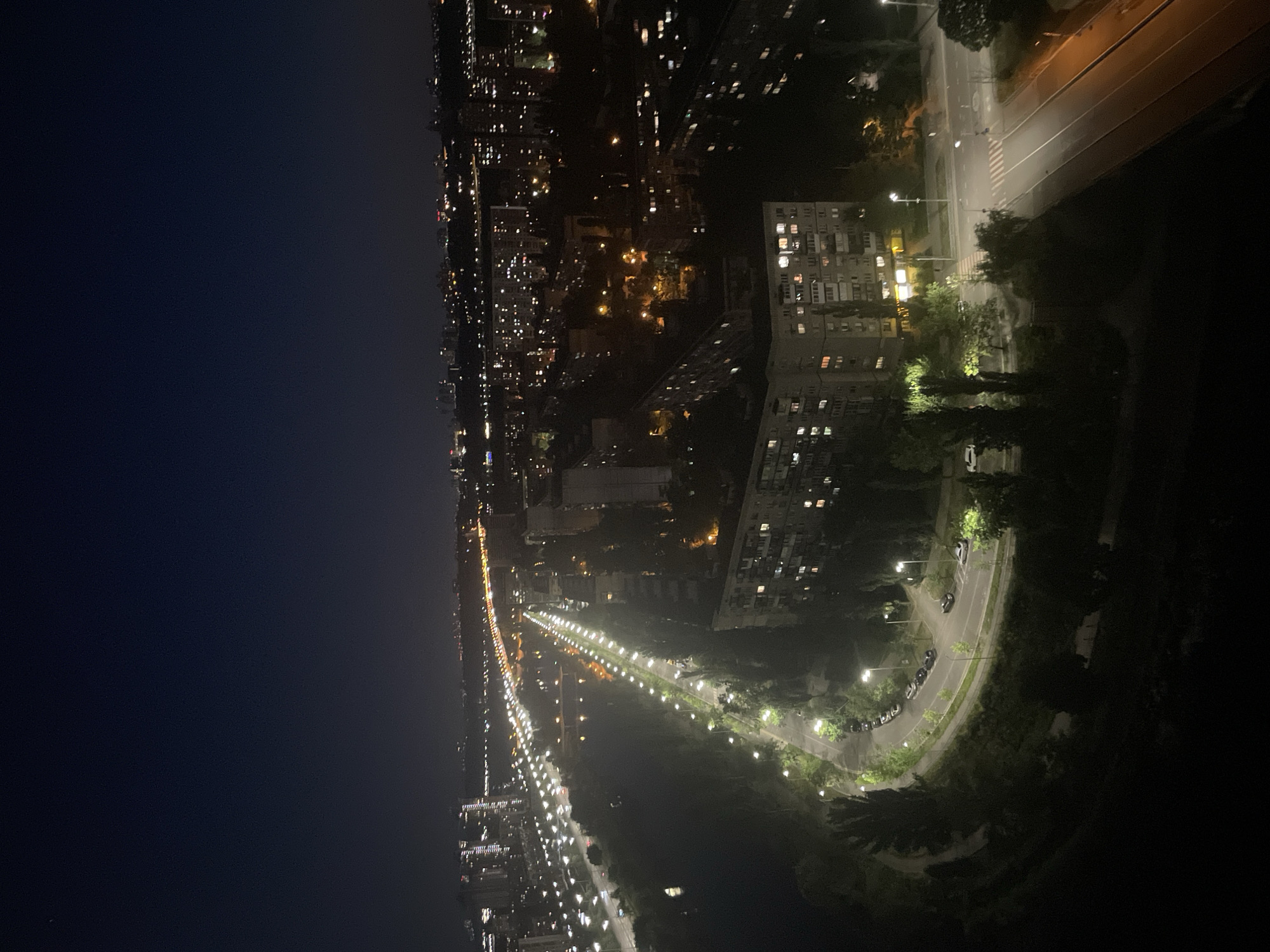 This is a nighttime aerial view of a city, likely in Kyiv, Ukraine, characterized by its illuminated streets, buildings, and a dark, expansive sky. A prominent feature is a wide, curving road lined with streetlights that casts a bright glow on the pavement. Several parked cars are visible along the side of the road. Adjacent to the road is a stretch of green space, possibly a park or a tree-lined embankment, with trees casting shadows. In the background, apartment buildings with illuminated windows are visible, suggesting a residential area. Further in the distance, a dense cityscape unfolds with numerous buildings and a highway or a long road illuminated by rows of streetlights, indicating a larger urban area. The overall impression is one of a vibrant city at night, with artificial lights providing the primary illumination against the dark natural background of the sky.