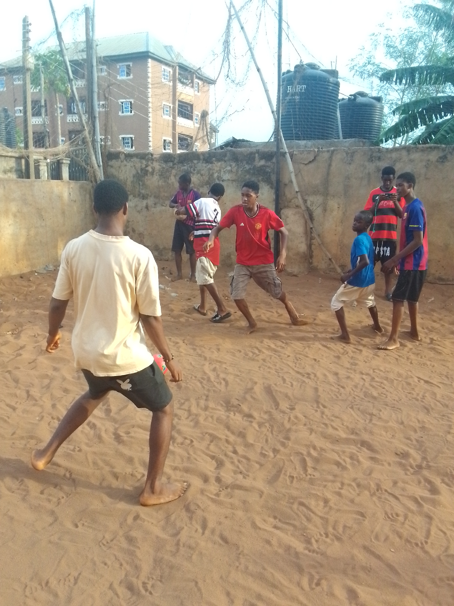 The image shows a group of young men playing soccer barefoot on a sandy ground, likely a recreational area, in the city of Unknown, Unknown. In the foreground, a player in a light-colored shirt and dark shorts faces away from the camera. Several other players are clustered near the center of the frame, wearing various jerseys and shorts. Behind them, a weathered concrete wall stretches across the scene. Above the wall are water tanks and some greenery, and in the background, a multi-story building is visible. The sky is overcast.