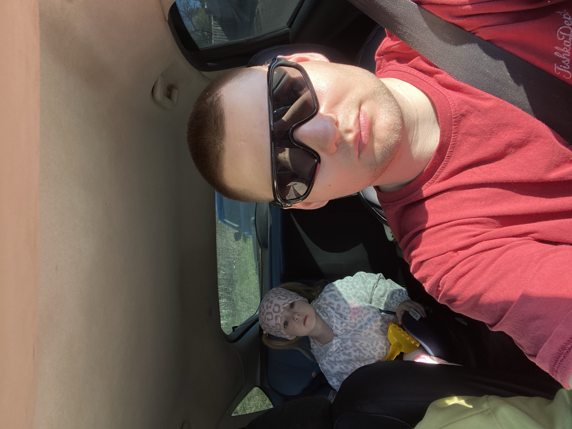 The image shows a man wearing sunglasses and a red t-shirt with a visible seatbelt. He is in the driver's seat of a car. A child is visible in the back seat, wearing a patterned hooded jacket and a headscarf. The child is holding a yellow object. The location is likely Chaikovsky, Russia, though this is not directly verifiable from the image.
