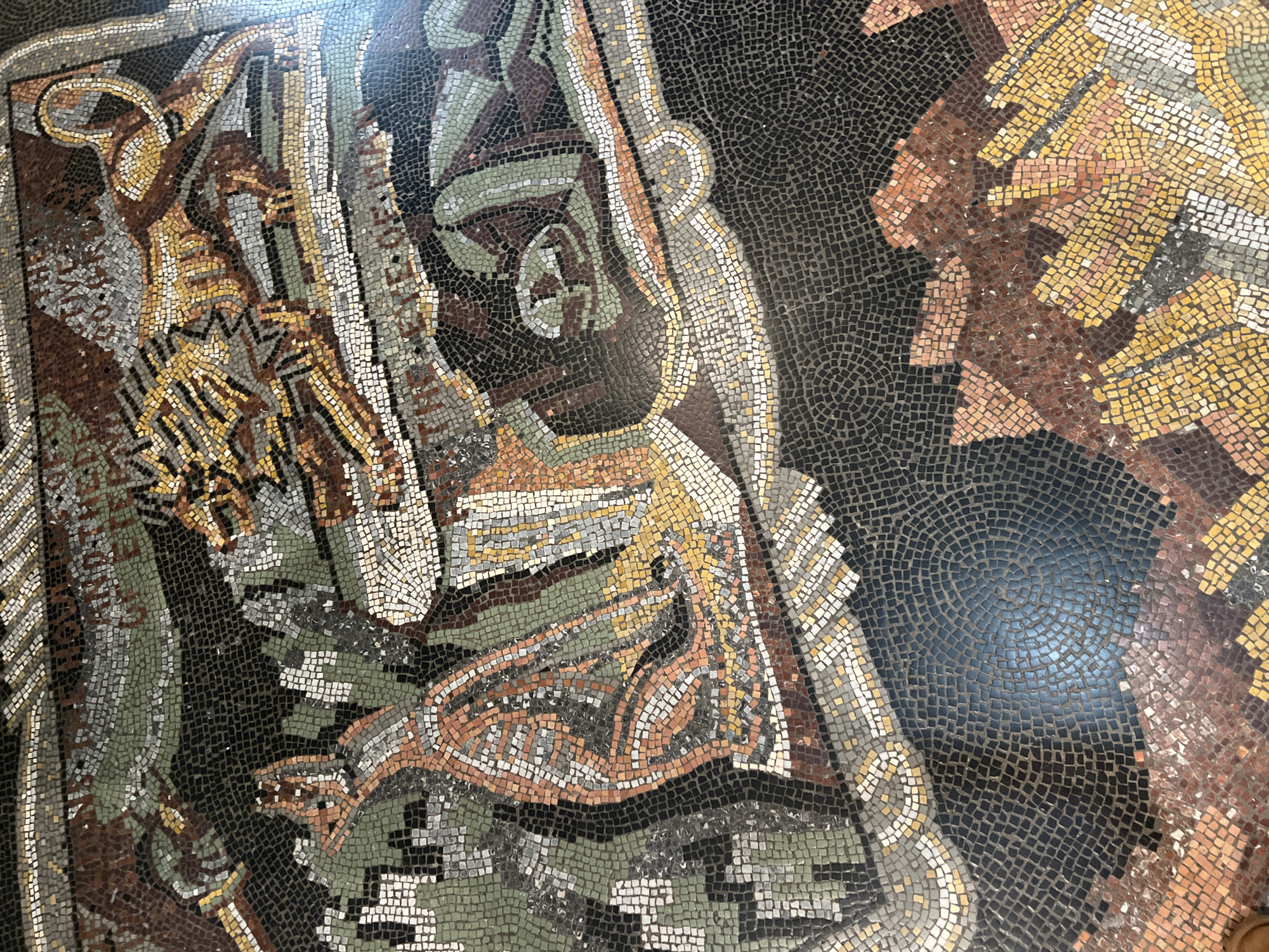 This media file presents a detailed, close-up view of an intricate mosaic, likely adorning a floor or wall. The artwork features a complex, stylized design composed of numerous small, irregularly shaped tesserae.
The dominant color palette includes earthy tones such as various shades of brown, gold, yellow, cream, and grey, accented by muted greens. These colors are set against a deep, almost black background, which makes the lighter elements stand out prominently. The tesserae exhibit subtle variations in hue and texture, contributing to the depth and handmade feel of the piece.
The design appears to be part of a larger, possibly figurative or mythological scene, though the full context is not visible. Within the frame, curvilinear forms and flowing patterns are prominent. One section shows a bold golden curve, possibly representing a stylized limb or an ornamental flourish. Other areas feature intricate patterns that could be interpreted as scales, feathers, or decorative motifs, created by the careful arrangement of different colored tesserae. There are also sections with more geometric, zigzag patterns, adding to the visual complexity.
The dark background tesserae, particularly visible in the crops, sometimes form subtle circular patterns, hinting at larger design elements beyond the immediate focus. The surface of the mosaic has a slight sheen, reflecting some ambient light. As a static artwork, no specific activity or event is taking place within the image. There is no visible text. The craftsmanship and material suggest an artwork rooted in historical mosaic traditions.