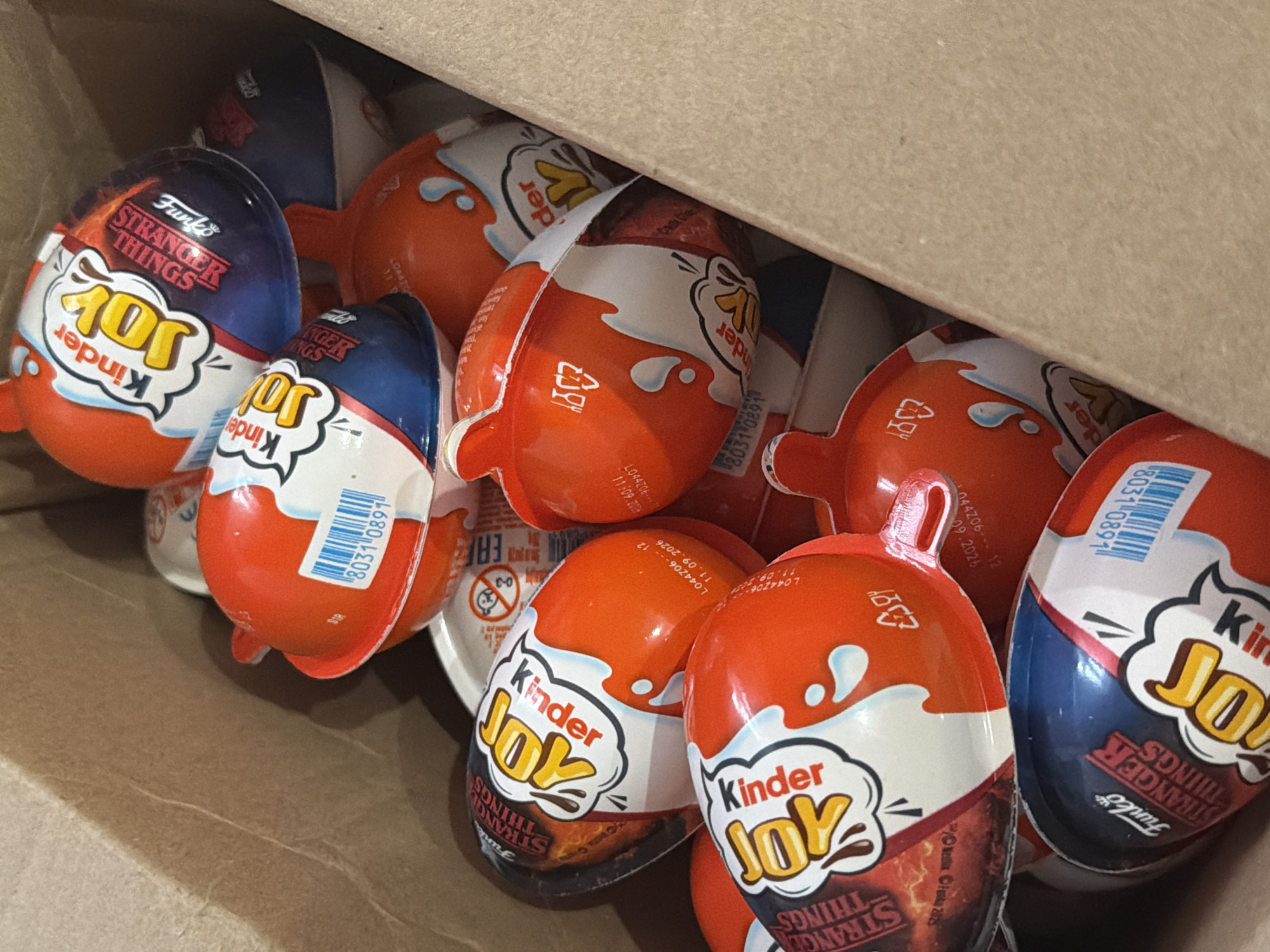 A close-up shot shows a pile of Kinder Joy eggs inside a brown cardboard box. The eggs are a mix of orange and navy blue, with "Kinder Joy" prominently displayed on the wrapper. Some of the eggs feature a "Funko Stranger Things" design. A barcode is visible on some of the eggs, reading "8031 0891". Expiration dates such as "11.09.2026" and manufacturing codes like "L044Z06--12" are also visible. The box suggests the presence of other eggs beneath the visible layer. No people are visible in the image. The location is not Perm, Russia, based on the available information.