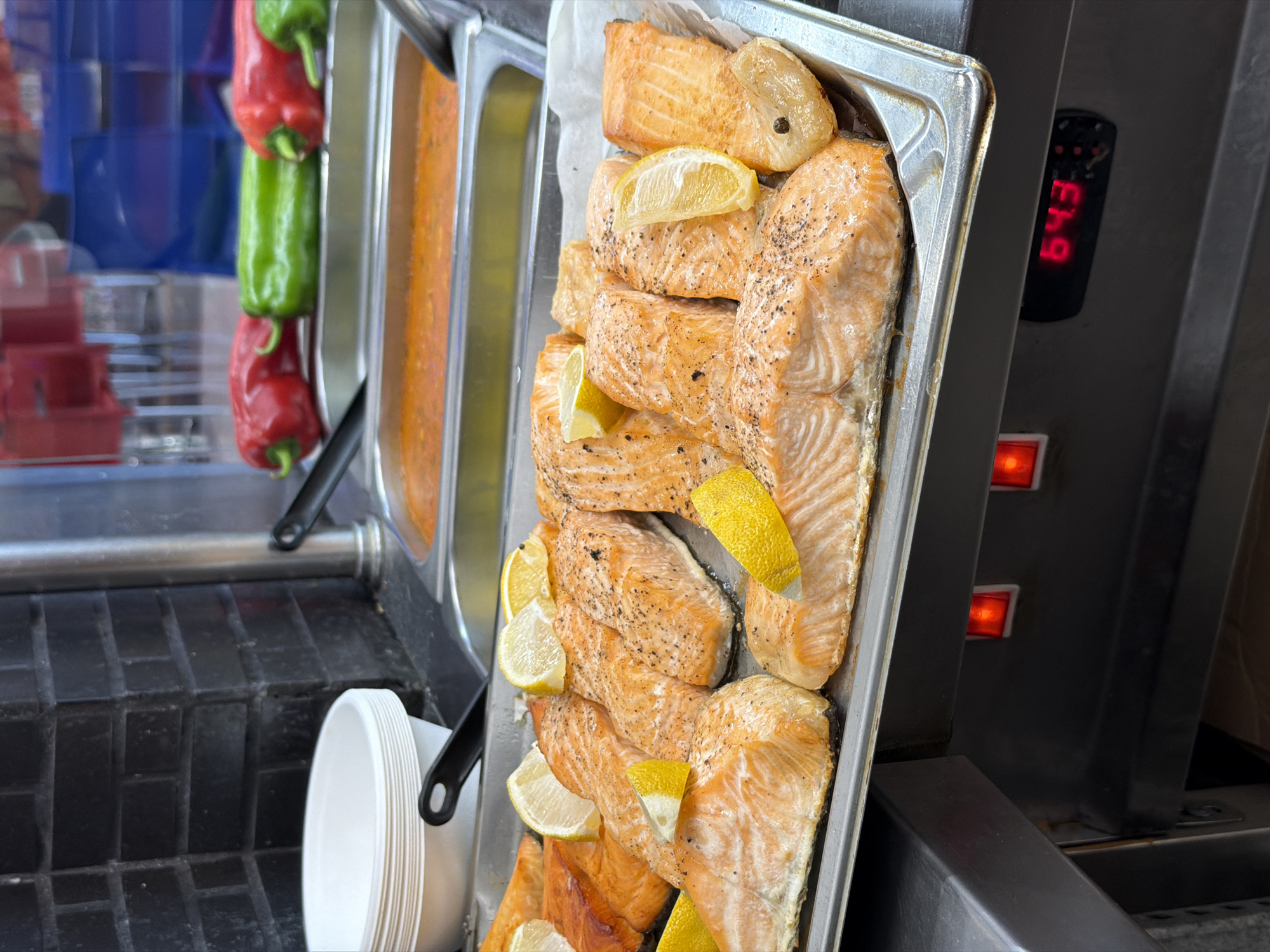 This image presents a detailed view of a food service counter, likely within a cafeteria or buffet setting, located in Plovdiv, Bulgaria. The foreground is dominated by a rectangular stainless steel tray containing numerous cooked salmon fillets. These fillets are light orange-pink in color, exhibit a flaky texture, and are visibly seasoned with dark specks, likely black pepper. Multiple lemon wedges are strategically placed atop and between the salmon pieces as garnish.
Directly behind the salmon tray, two additional large stainless steel inserts are visible, each holding a different prepared food item. The insert on the right contains a thick, reddish-orange liquid, suggestive of a sauce or stew, with a serving utensil partially submerged. The insert to its left contains a lighter orange liquid, also appearing to be a sauce or soup.
Above these liquid-filled inserts, on a slightly elevated shelf, are several whole bell peppers: two vibrant red peppers positioned on the left and two green peppers on the right.
To the far left of the frame, a stack of white, possibly disposable, serving bowls is present. A black-handled serving utensil, resembling a spatula, rests beside these bowls. The background on the left side features a dark, brick-patterned wall.
Below the food display area, the stainless steel counter reveals operational elements. Two illuminated red indicator lights are visible, indicating active power or heating. Centrally positioned below the food trays is a small black digital display module exhibiting the red numerical reading "64.3," which likely represents the holding temperature of the food, possibly in degrees Celsius. The overall background is blurred, showing indistinct reflections of an indoor environment, including what appear to be red and blue chairs, but no individuals are clearly discernible.