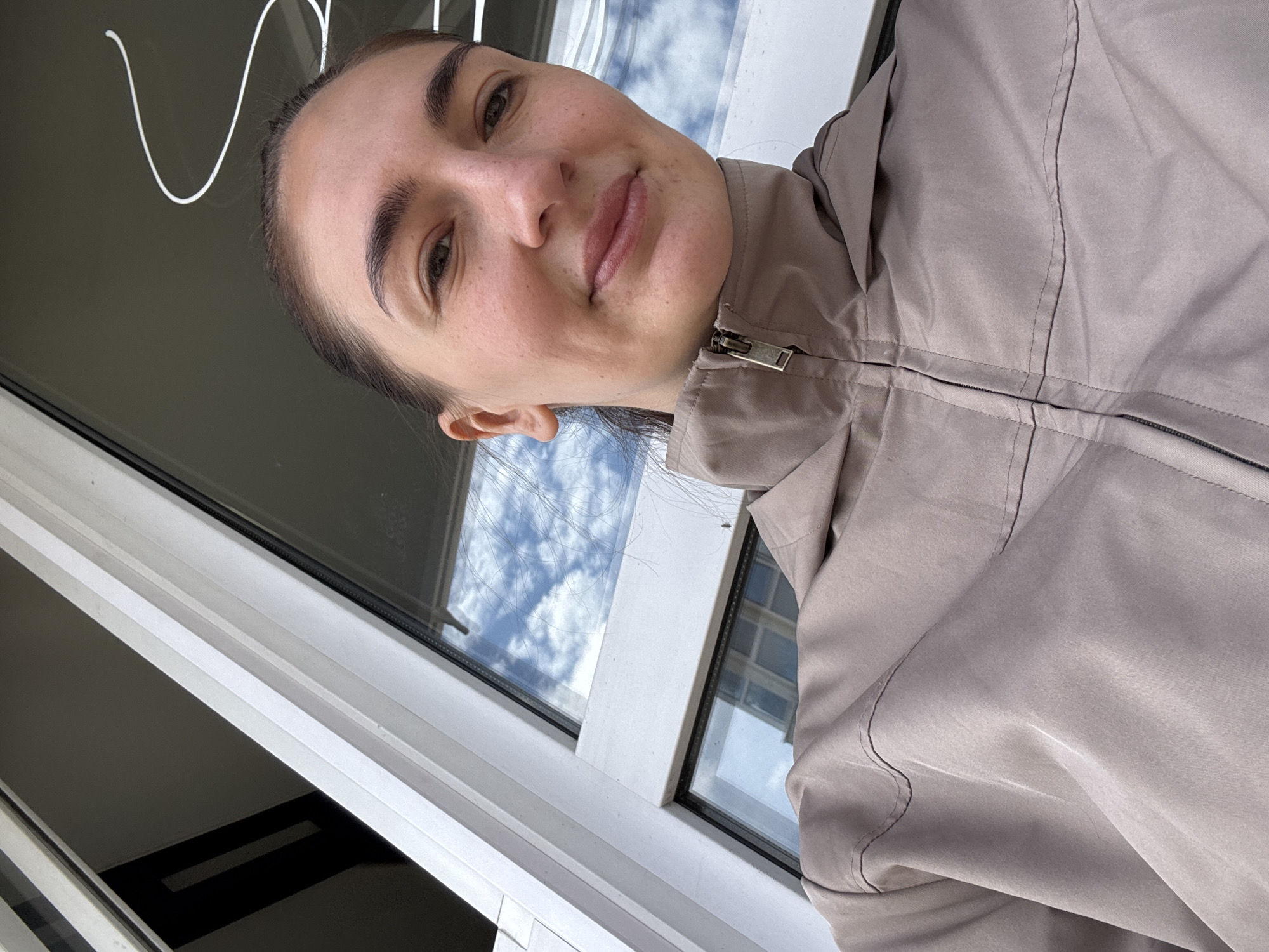 A woman with dark hair and light skin stands indoors near a window, wearing a tan zip-up jacket. The window reflects a cloudy sky. The location cannot be determined from the image.