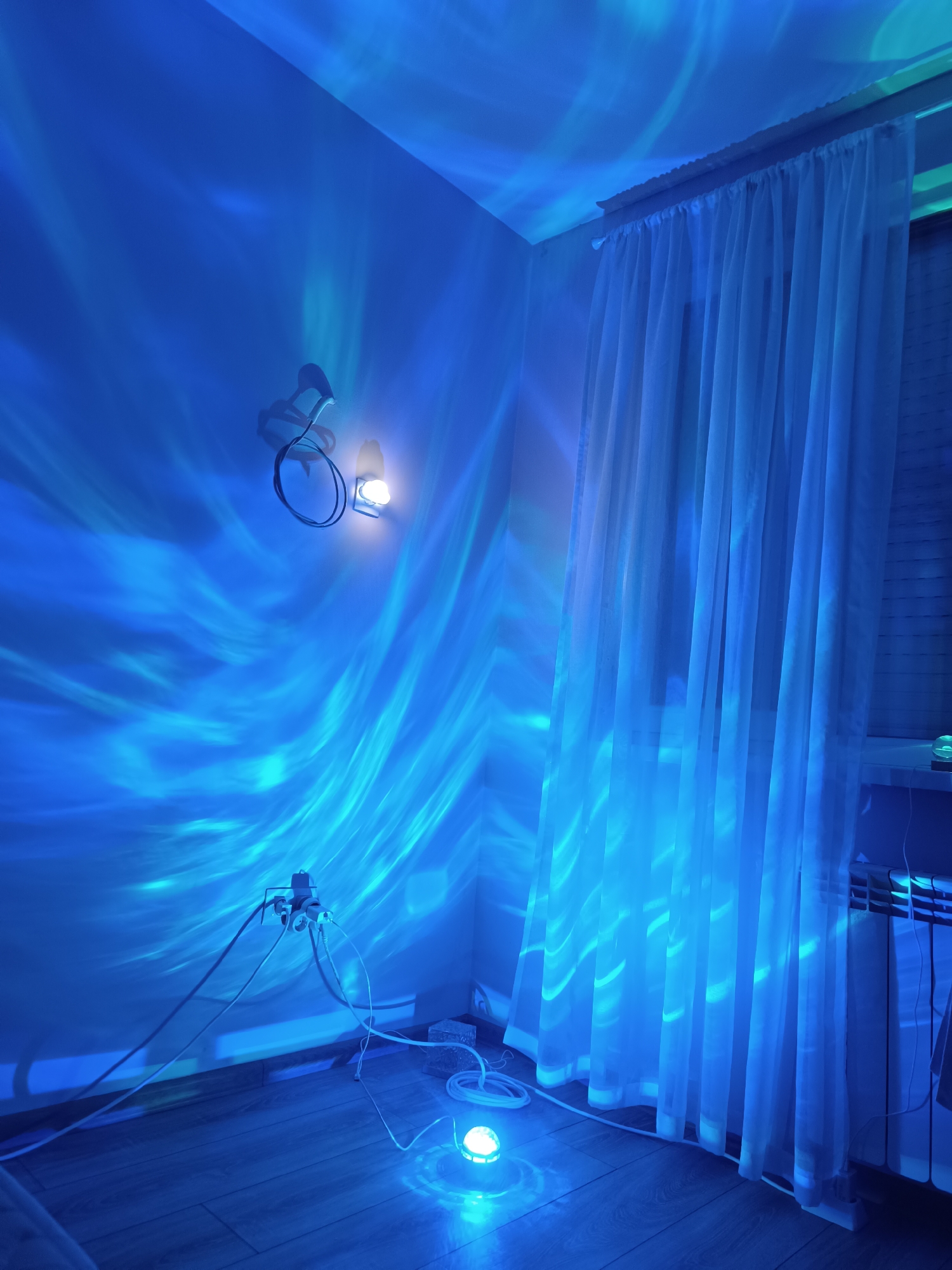 The image depicts an interior scene illuminated by blue light projections in a room.
**Location:** There is no indication of the location being Cherkovitsa, Bulgaria, based on the information present in the image.
**Environment:** The room features light hardwood flooring and white walls. The light projections create wave-like patterns across the walls and ceiling. A window with sheer white curtains is visible in the right side of the image.
**Objects:** A small spherical lamp sits on the floor, producing the blue light effect. An electrical outlet with multiple plugs and associated power cords are attached to the wall. A light fixture is mounted on the wall. There is a radiator.
**Actions/Interactions:** The lamp is projecting light, creating the wave-like patterns.