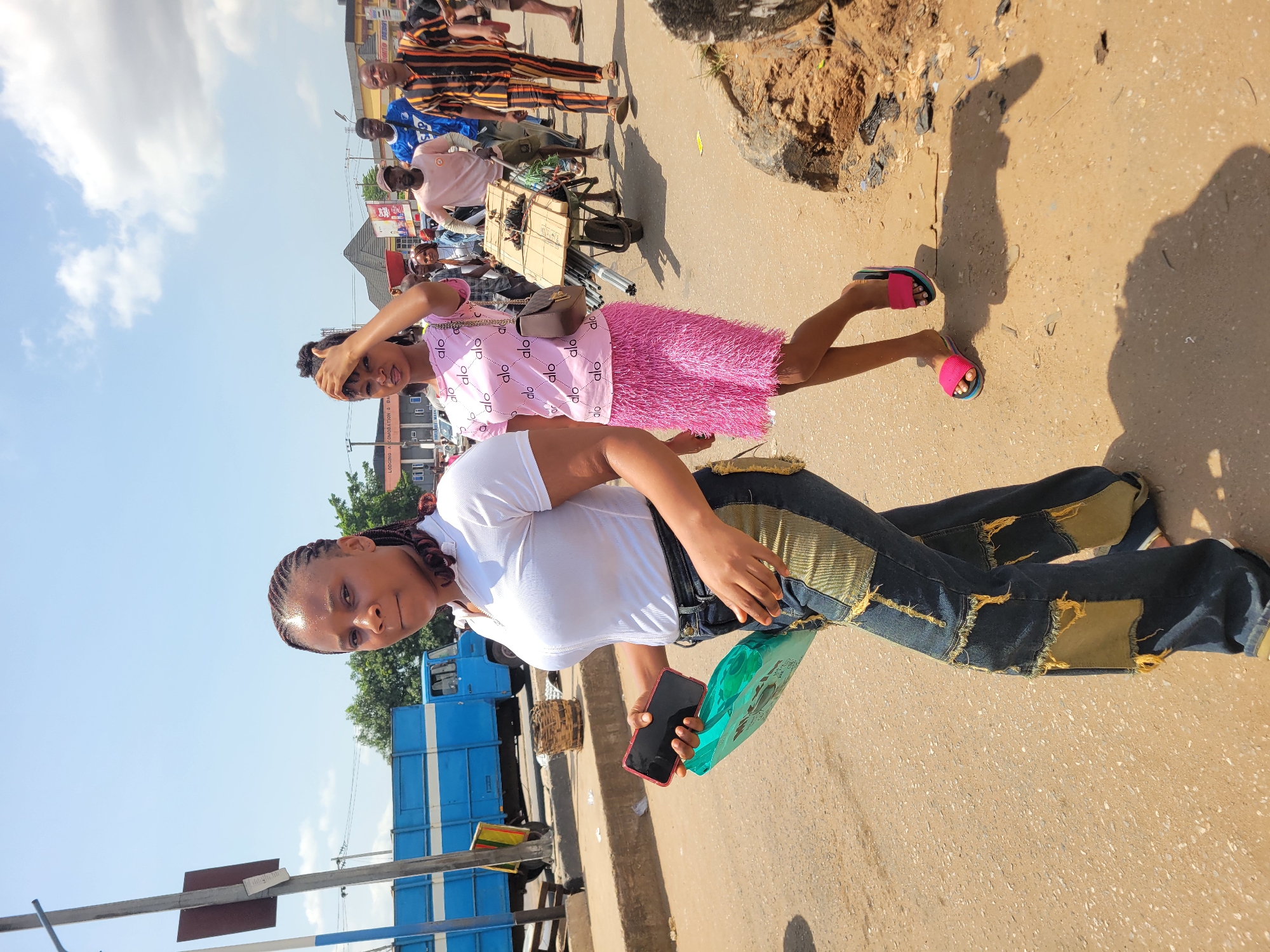 This media file captures a vibrant street scene in Onitsha, Nigeria, under bright midday sun. The image is primarily focused on two women in the foreground, with a bustling urban environment stretching into the background.
The woman closest to the viewer, positioned on the left, wears a white ribbed t-shirt and unique blue denim jeans featuring distinct yellow and green patches, some with frayed edges. Her hair is styled in dark braids. She holds a red smartphone in her right hand and a translucent green object, possibly a bottle or small bag, in her left, looking directly towards the camera with a neutral expression.
Behind her and to the right, another woman is partially visible. She wears a striking pink dress adorned with repeated "alo" patterns and pink flat sandals. One hand is raised towards her head, possibly shielding her eyes from the bright sunlight or adjusting her hair. Her face is turned away from the camera.
The setting is a busy street, likely a market area, with several other pedestrians visible in the background, some walking and others appearing to be vendors with pushcarts. A large blue truck is parked on the left side of the street, and various buildings and street infrastructure can be seen further back. The ground is a mix of asphalt and dirt, with visible strong shadows cast by the intense sunlight, suggesting clear weather. Visible text includes "LODGING A COMODATION & BA" on a distant sign and the "alo" pattern on the pink dress.