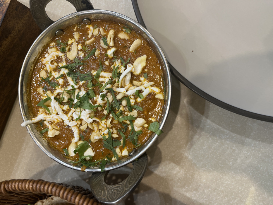 A metal bowl filled with a brown gravy, topped with shredded white cheese, cashews, and chopped green parsley, sits on a beige countertop. The bowl has a decorative handle. A white plate, with a brown rim, is to the right of the bowl. The scene is in Sahibzada Ajit Singh Nagar, Punjab, India. The location is based on the coordinates: latitude 30.718, longitude 76.714.