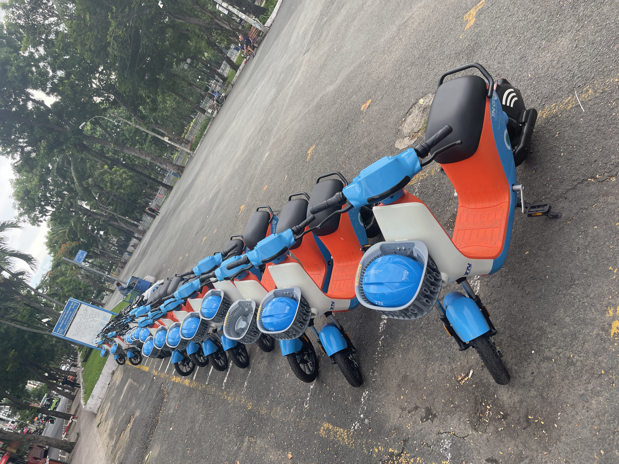 The image depicts a row of electric scooters parked on a paved surface, likely a street or parking area, in Thủ Đức, Vietnam. The scooters are predominantly orange and blue, with a white section on the front. They have black seats and handlebars, and some are equipped with blue helmets in front baskets. The scooters are arranged in a line, angled towards the right side of the frame. In the background, a line of trees with lush green foliage provides shade. The ground is asphalt, with yellow markings indicating parking spots. There are some leaves scattered on the ground.
In the distance, a few people can be seen, including individuals on bicycles and a person sitting on a bench. A signpost with Vietnamese text and a map-like diagram is visible on the left side of the image, suggesting this is a designated parking or rental area for the scooters. The overall scene appears to be during the daytime, with overcast or diffused lighting, possibly indicating a cloudy day or early morning/late afternoon. The presence of trees and the general ambiance suggest an outdoor urban or suburban setting.
The visible text on the scooters includes "TNG GO" and some green circular logos. The signpost also contains Vietnamese text and diagrams, providing information about the area or the scooter service. There are no strong indicators of specific emotions, but the organized parking of the scooters suggests a functional setup for transportation or rental services. The overall impression is one of a modern, convenient urban environment offering electric scooter options.