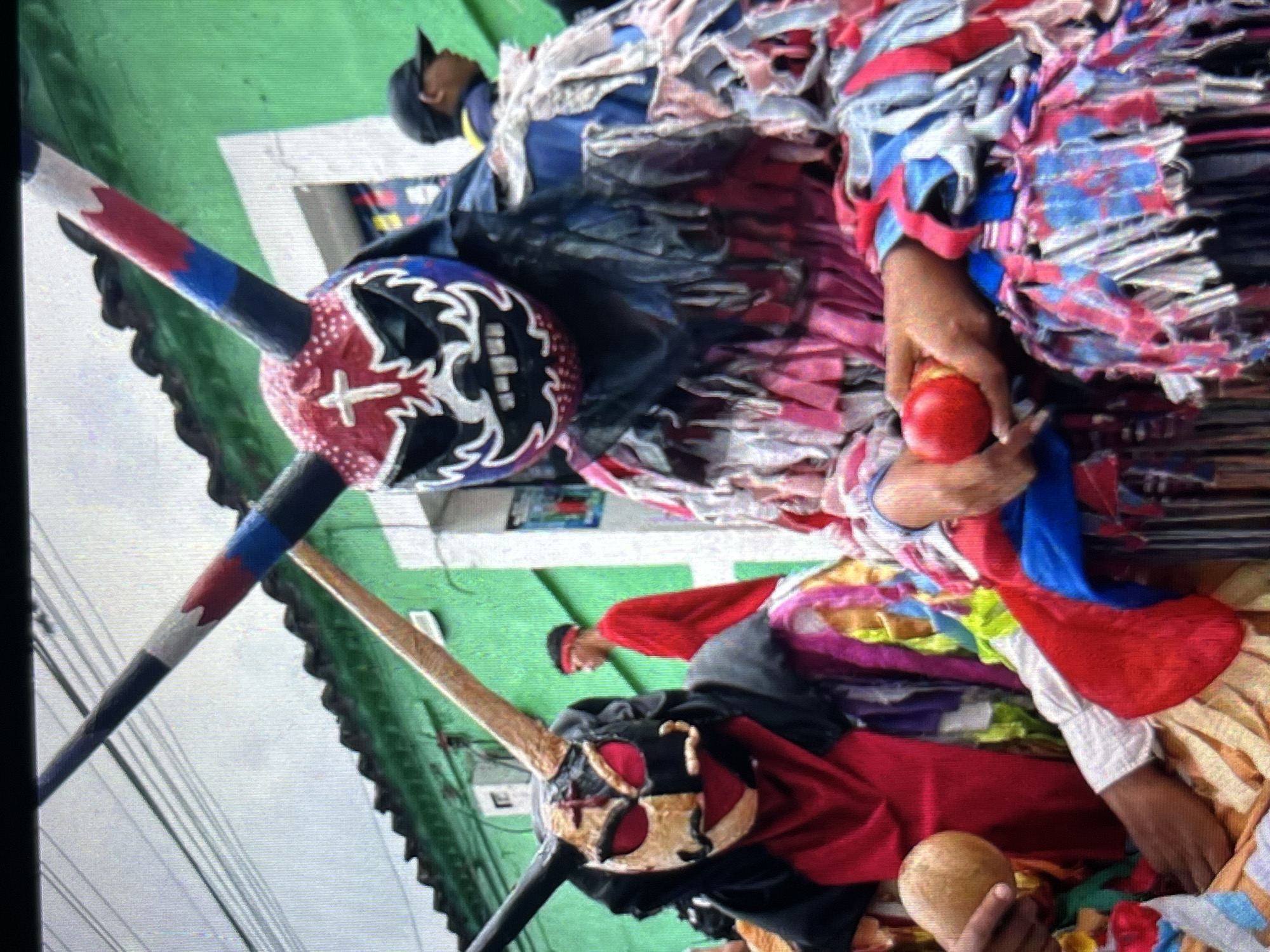 In Tinaquillo, Venezuela, a street scene is depicted featuring individuals in elaborate, traditional costumes. The foreground is dominated by two figures wearing masks and ornate, fringed garments. The mask on the right is red, blue, and white with large horns painted black, red, and blue, featuring a cross on the forehead and sharp teeth. This figure wears a black fringe headdress and a pink and multi-colored fringed costume. Their right hand is holding a red apple. The mask on the left is split in two colors, black and tan, with red eyeholes and also features horns, this one appearing more rustic and golden-brown. This figure wears a red garment underneath their black and colorful fringed costume and holds a gourd-like object in their left hand. A person wearing a red shirt and a red headband is visible in the background, along with other individuals in similar festive attire and a person wearing a black baseball cap. The background consists of a green building with a tiled roof, suggesting an outdoor public space in the town. Overhead, power lines are visible against a bright sky.