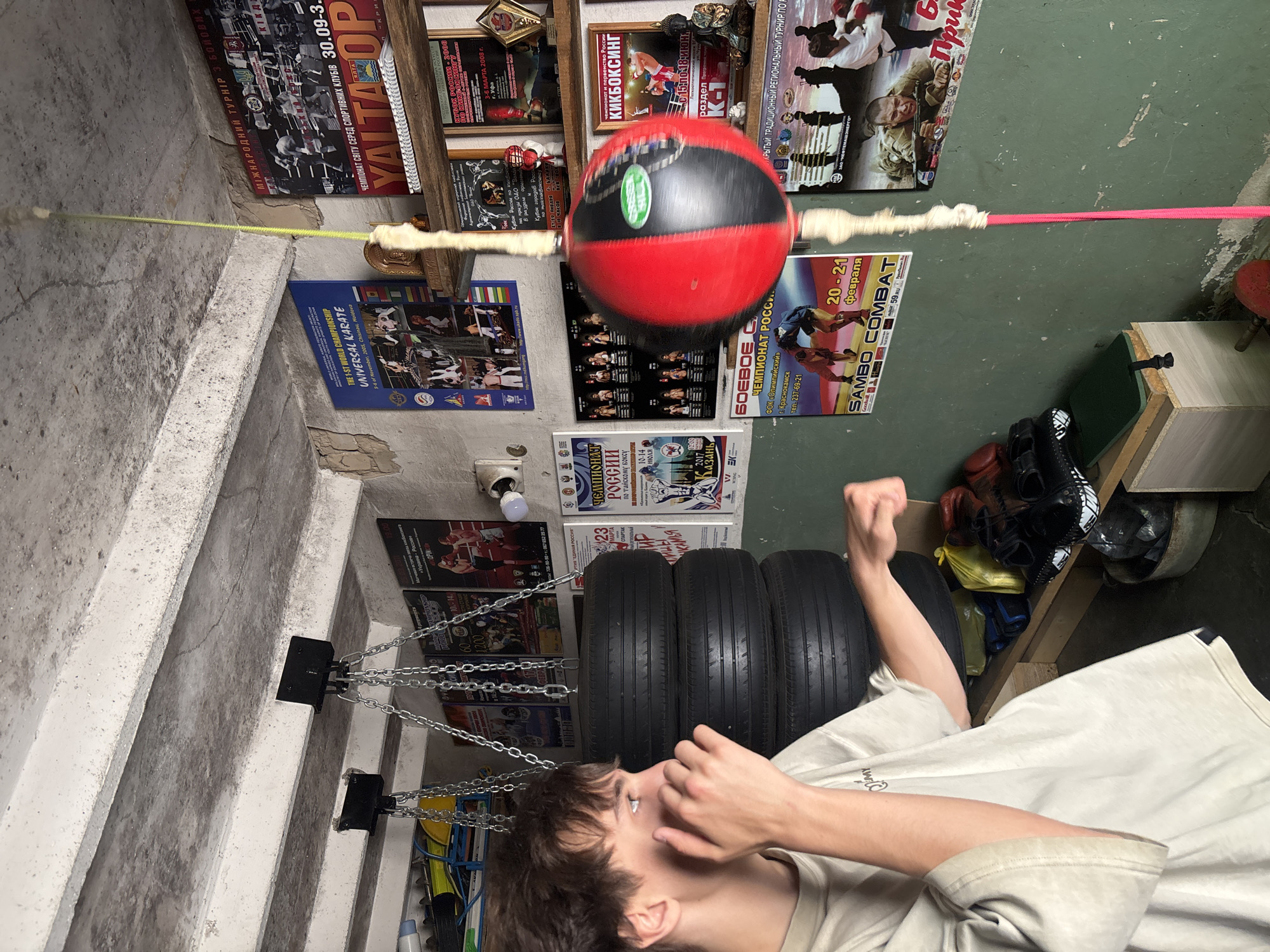 A person is in a boxing training space, likely a basement, possibly in Chaikovsky, Russia based on the numerous posters on the walls including "SAMBO COMBAT". They are striking a hanging red and black speed bag. The space has a concrete wall, stairway, and floor. Tires hang suspended by chains. Additional boxing gear and posters are mounted on the wall. The person appears to be wearing a t-shirt. The room is dimly lit.