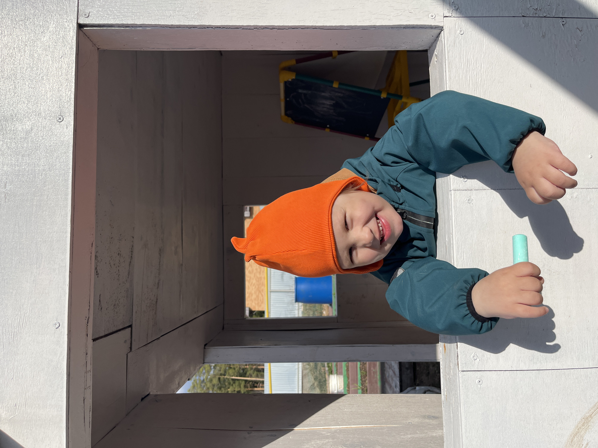 A young child, wearing a vibrant orange beanie and a teal jacket, smiles broadly while peeking out from a rectangular opening in a white wooden structure. The child's left hand holds a piece of light blue chalk, resting on the white frame of the opening. Their other hand is also visible, gripping the opposite side of the frame.
The setting appears to be outdoors on a bright, sunny day, evidenced by the strong shadows cast on the white wooden surfaces. In the background, through the opening, a black easel or chalkboard with a yellow frame is partially visible, suggesting a play area. Further back, beyond the immediate structure, other buildings, possibly residential or utility, including one with a blue barrel, and some trees can be seen, indicating a suburban or rural outdoor environment. The interior walls of the wooden structure are also white-paneled. The child's expression is one of happiness and joy. No visible text is present in the image. The location context for this scene is Chaikovsky, Russia.