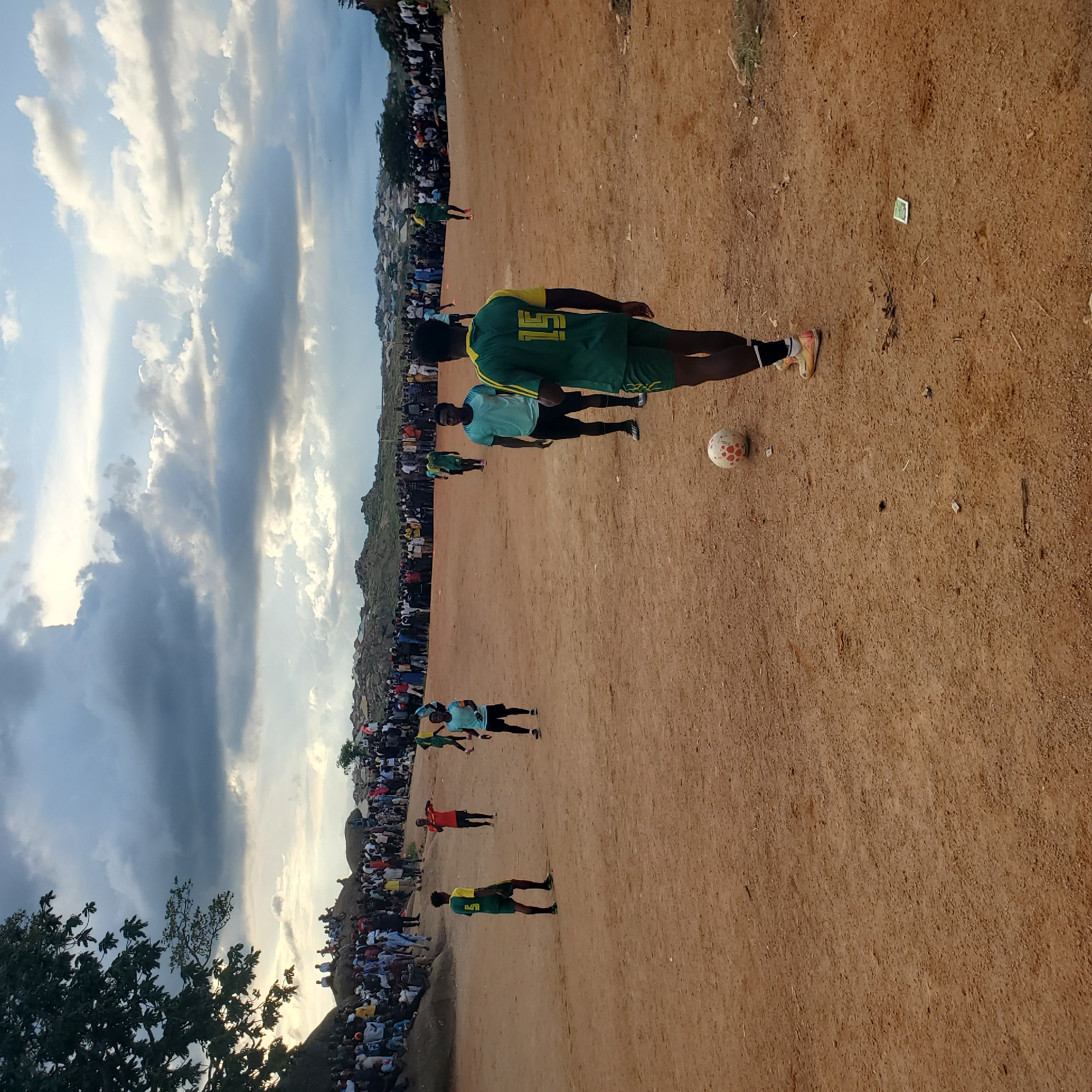 An outdoor soccer match is underway on a dirt field in Jos, Nigeria. A crowd of spectators lines the perimeter of the field, some seated on rocky outcrops and others standing. Several players in green and yellow uniforms are visible on the field, as well as one player in a light blue uniform and another in a red uniform. A soccer ball is present on the field. The sky is partly cloudy. The area appears to be a residential neighborhood with buildings in the background.