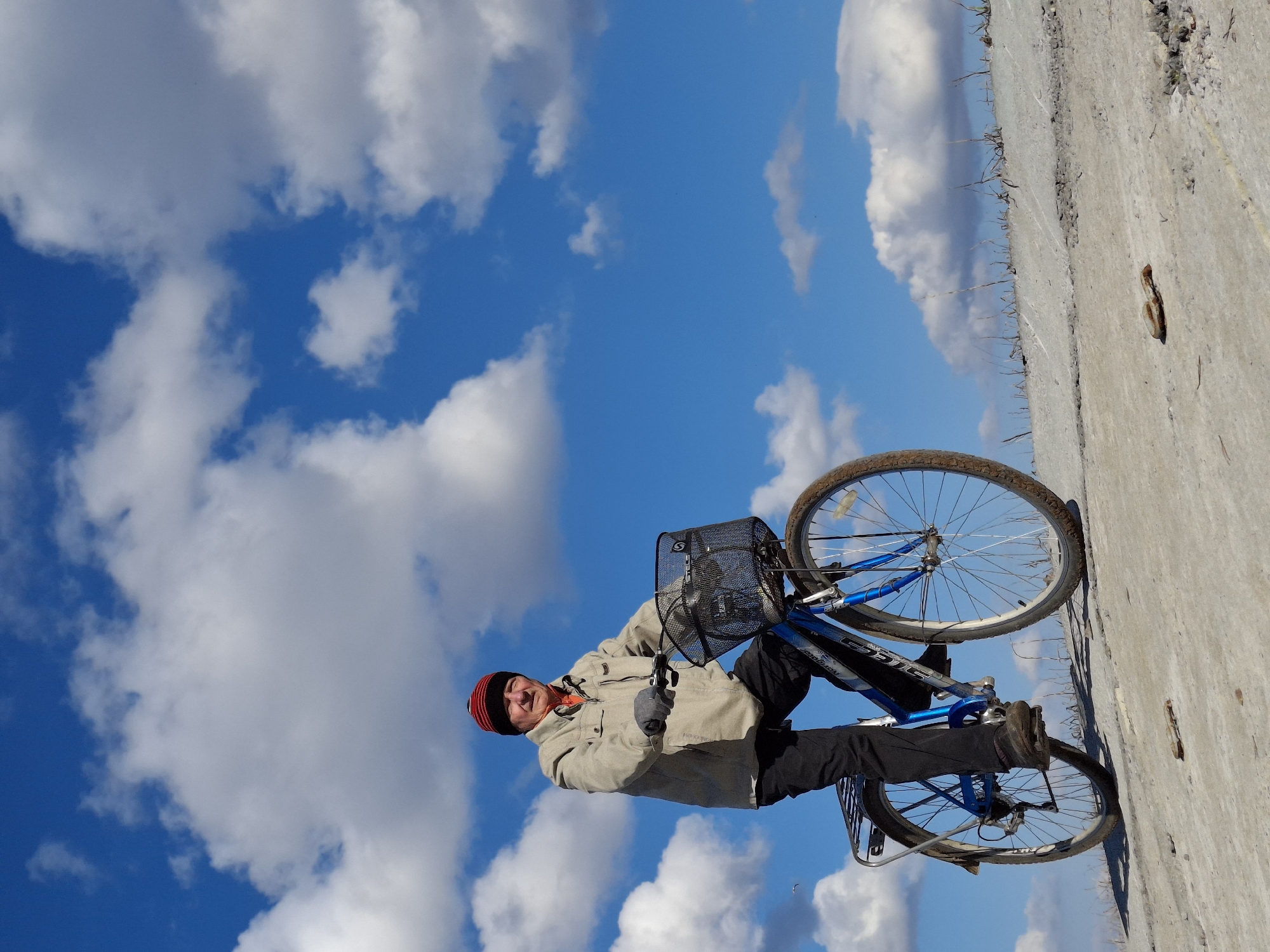 The image portrays an older man standing outdoors with his bicycle under a bright blue sky dotted with large, fluffy white clouds. The man, positioned on the left side of the frame, is wearing a red and black knit hat, a light beige or off-white jacket over an orange top, black pants, and dark gloves. He has a pleasant, slightly smiling expression.
He is holding onto a blue bicycle equipped with a black mesh front basket, which displays an "S" logo, and a rear luggage rack. Faint text visible on the bike's frame includes "NAVIGATOR" and "STELS".
The setting appears to be a concrete or paved path, possibly alongside a larger concrete structure like an embankment or wall on the right. Sparse, dry vegetation or weeds are visible growing along the edge of the paved surface. The scene is bathed in clear daylight, indicating sunny weather.
The activity depicted is the man pausing with his bicycle, suggesting a break from a ride or posing for a picture in a relaxed moment. The location context for this scene is Chaikovsky, Russia.