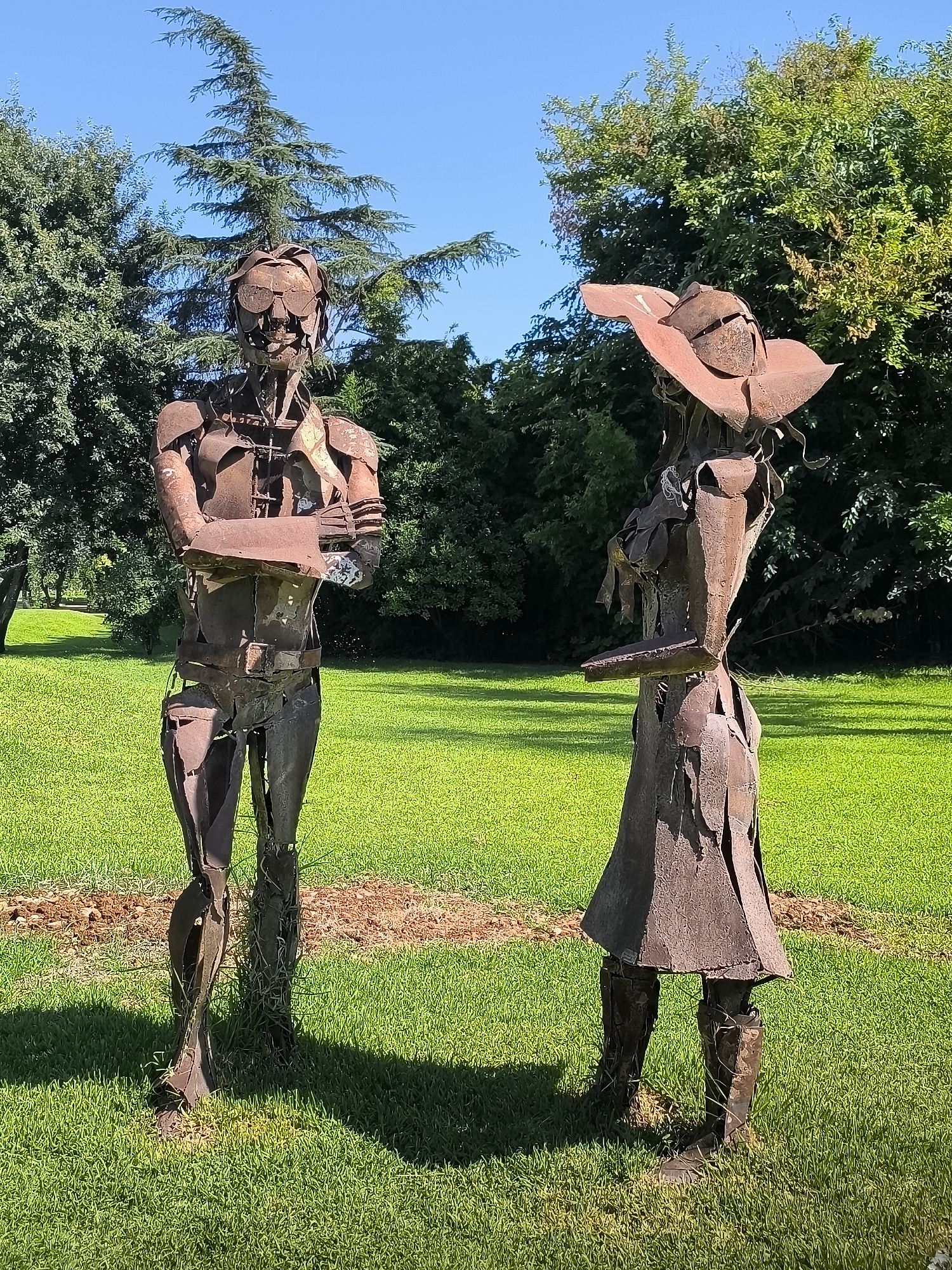 The image displays two full-body sculptures made of rusted metal, positioned in a grassy outdoor area. The sculpture on the left depicts a standing figure with arms crossed, appearing somewhat skeletal. The sculpture on the right is slightly shorter, wearing what resembles a hat and a skirt. Both sculptures have a rough, abstract form. The background consists of lush green grass, trees, and a clear blue sky. There is no indication that this scene is located in the city of Antalya, Turkey.