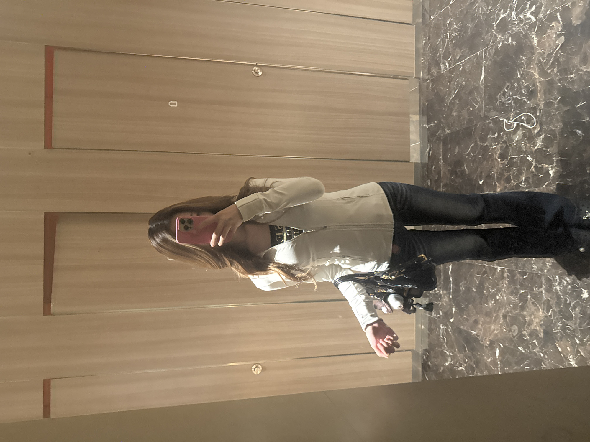 The image shows a person standing in what appears to be a restroom or similar enclosed space, taking a selfie in a mirror. The person is wearing a light-colored jacket, dark jeans, and dark shoes. They are holding a pink phone in front of their face. A purse or bag is hanging from their side. The environment consists of a wall with horizontal wooden panels and a marble-like floor or wall. It's impossible to determine if this image was taken in Yuzhong District, China, without further information.