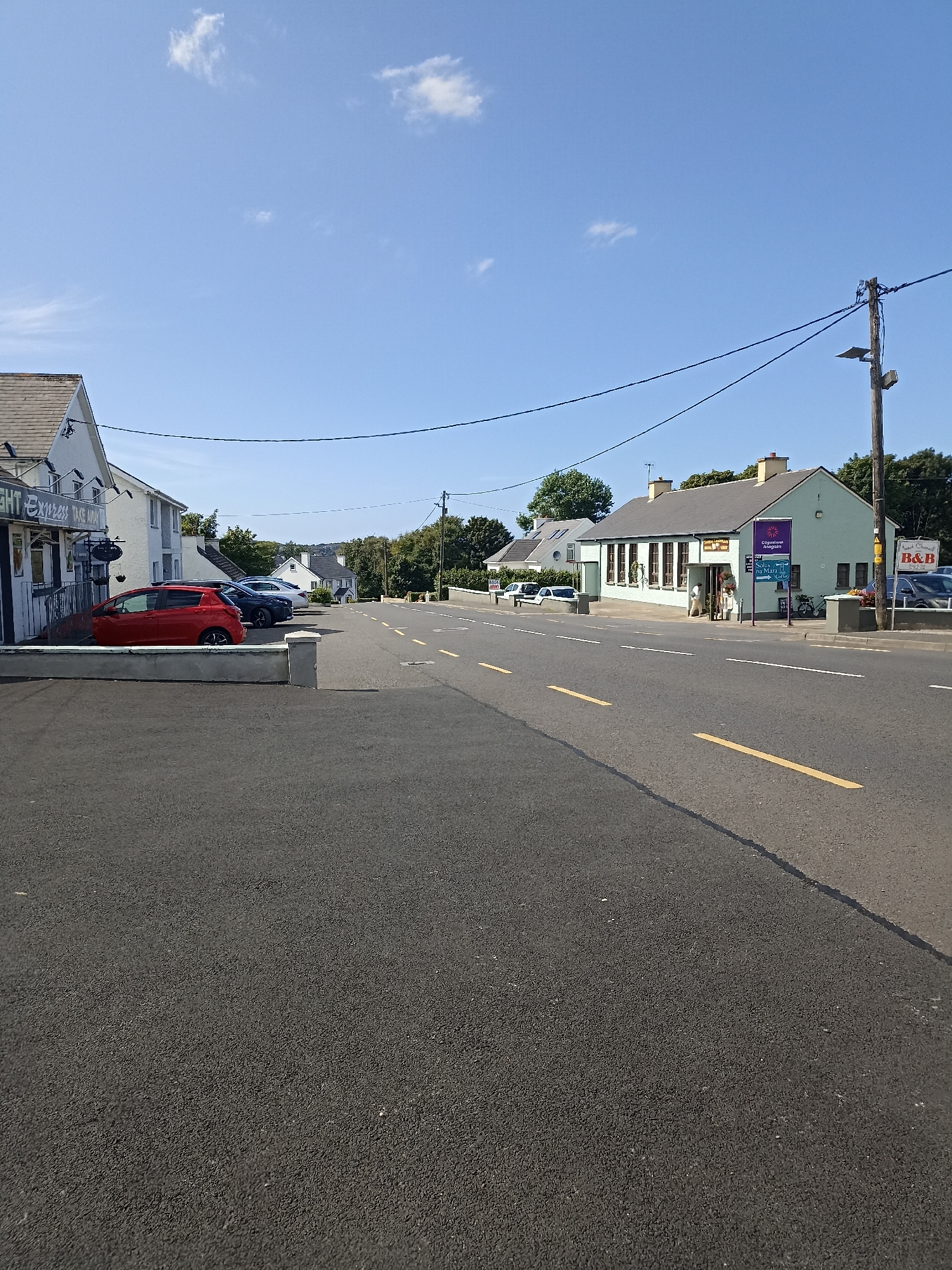 The image shows a street scene in Annagry, Ireland. A road with yellow dashed lines runs through the center of the frame, with businesses and buildings lining one side. On the left, a building with a sign that reads "LIGHT EXPRESS TAKE AWAY" is visible, with a red car parked in front of it. Further down the street, there is a light green building with a sign indicating "B&B" and another sign with Irish text, which translates to "Connemara Alehouse" and "Tea Rooms". Several parked cars are present along the street. The sky is clear and blue with a few scattered clouds. The environment appears to be a typical small town street with a mix of commercial and residential buildings.