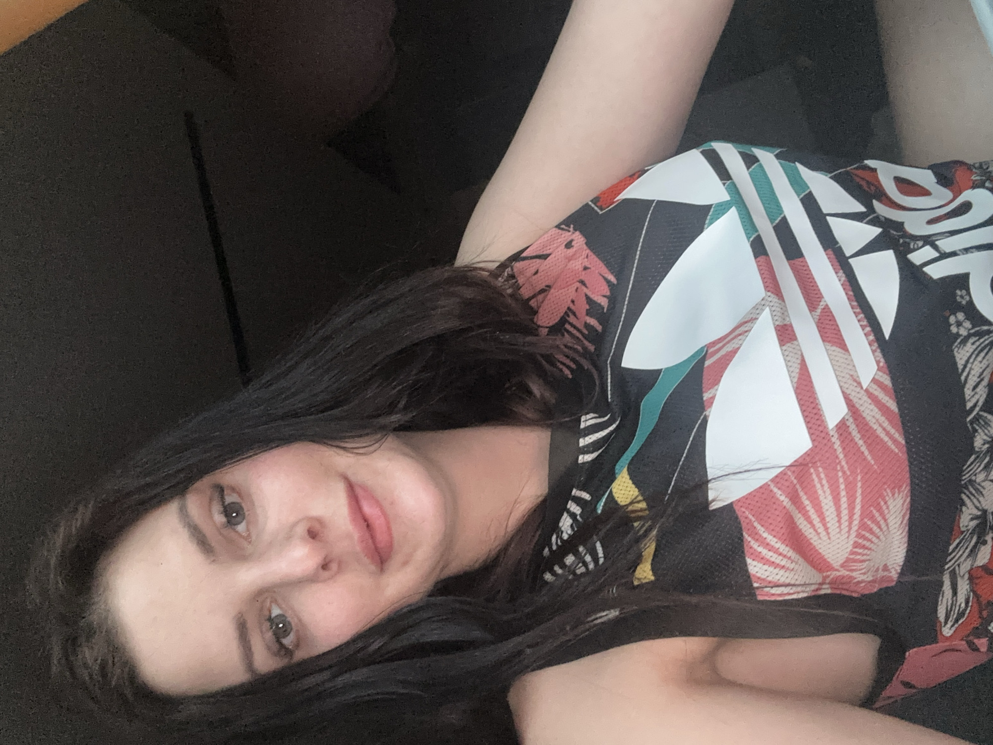 A woman is pictured from the chest up, looking directly at the camera. She has dark brown hair that falls past her shoulders and is wearing a black tank top with a colorful Adidas logo and a tropical print. The background is dark and appears to be indoors.
