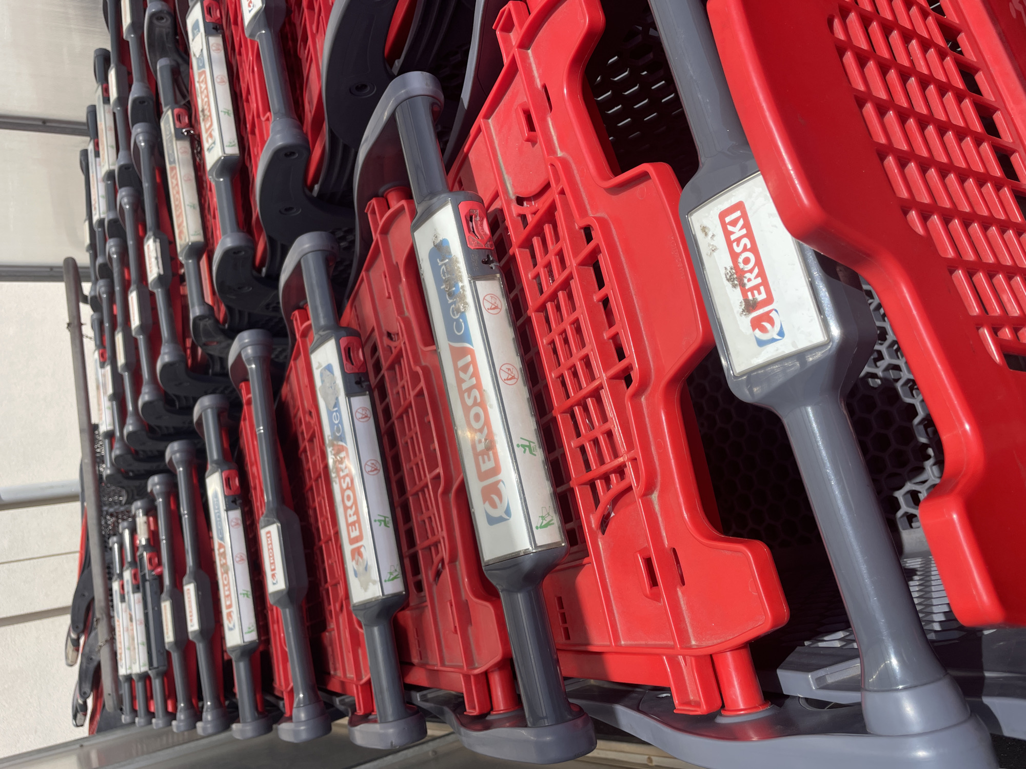 A line of stacked red and gray shopping carts is visible in what appears to be an outdoor commercial area in Gibraltar, Gibraltar. The carts are arranged in a nested formation, with their handles extending outward. Each visible cart has a "EROSKI center" label on its handle, with a logo featuring a stylized 'Q' and the word "EROSKI" in red. The handles also display icons of a shopping cart and a person with an outstretched arm, suggesting accessibility features. Some of the labels show signs of wear and tear. The carts are made of plastic and metal, with the baskets being a bright red and the handles a dark gray. The background is out of focus but suggests a building with vertical panels, possibly a retail store or a parking structure. No individuals are present in the frame. The lighting suggests it is daytime.