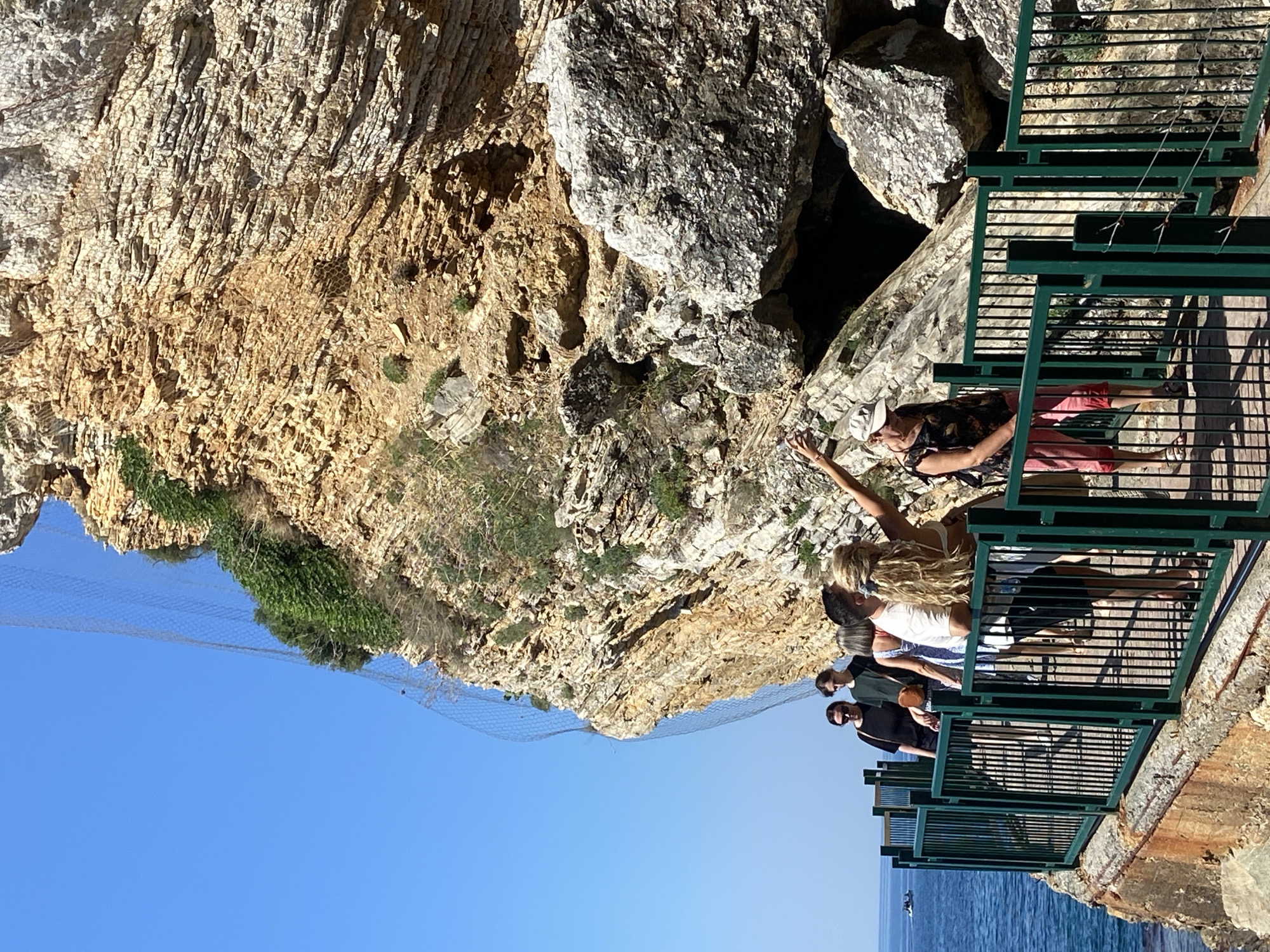 In Budva, Montenegro, a group of people stand on a scenic viewing platform overlooking a rocky cliff face and the shimmering blue sea. The platform, constructed with green metal railings, offers a vantage point to admire the natural landscape. A large net is stretched across the cliff, likely a protective measure against falling rocks.
The subjects include several individuals, some of whom are looking out at the view. One woman, with blonde hair, is reaching out towards the cliff, possibly pointing something out or gesturing. Another woman, wearing a hat and a patterned shirt, is also on the platform. The sky is a clear, vibrant blue, suggesting a sunny day. The sun appears to be high in the sky, indicating it is likely midday. The overall mood seems relaxed and appreciative of the beautiful surroundings. There is no visible text in the image.