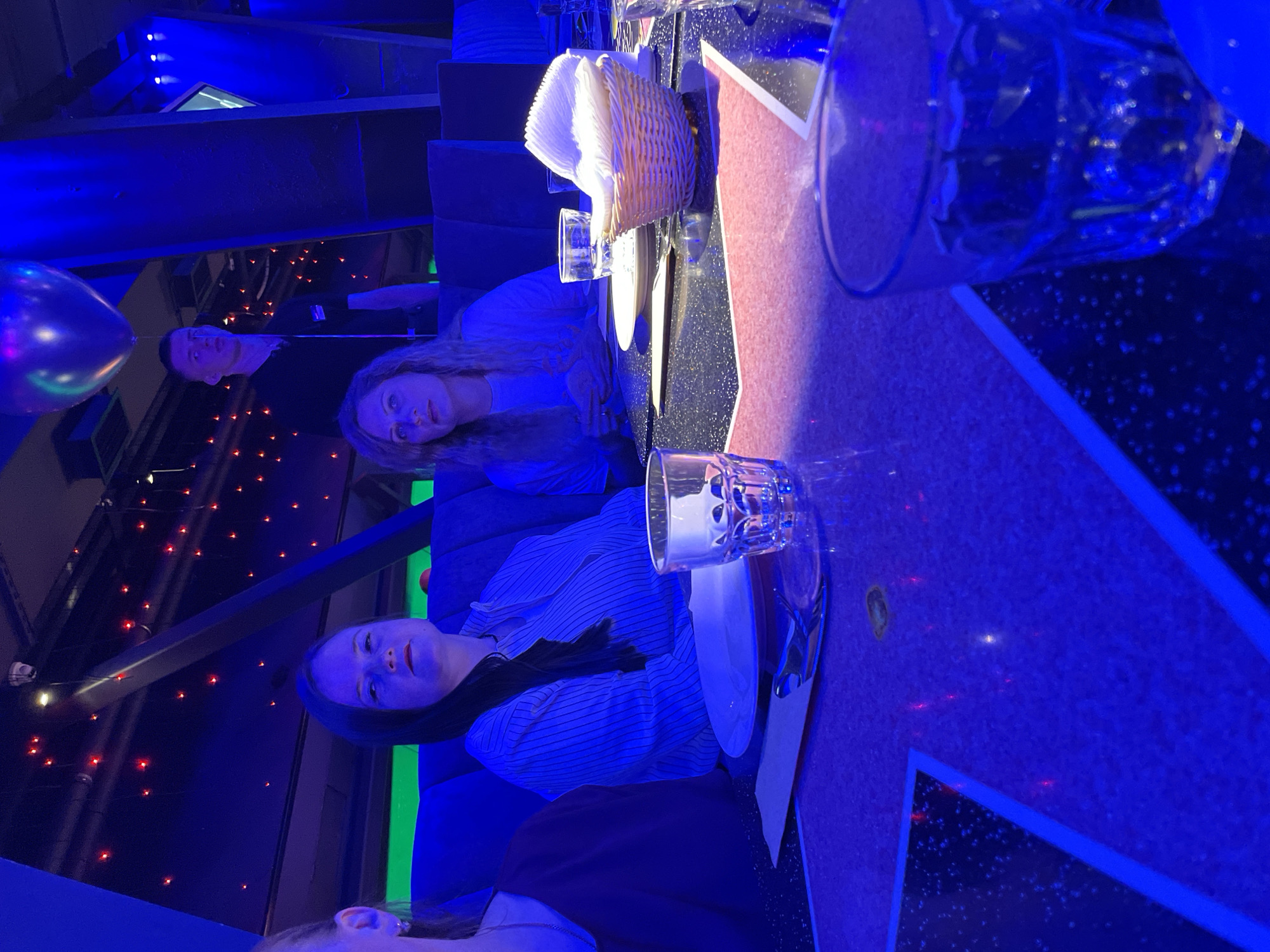A dimly lit restaurant scene in Saint Petersburg, Russia, features four individuals seated at a table. The table is decorated with plates, cutlery, glasses, and a basket of napkins. The ambient lighting is predominantly blue, with some red accents in the background. One person is visible on the far left, a second person is wearing a striped shirt, a third is wearing a light-colored shirt, and the fourth person's face is visible in the background, near a purple balloon. The table has a star-shaped design in blue.