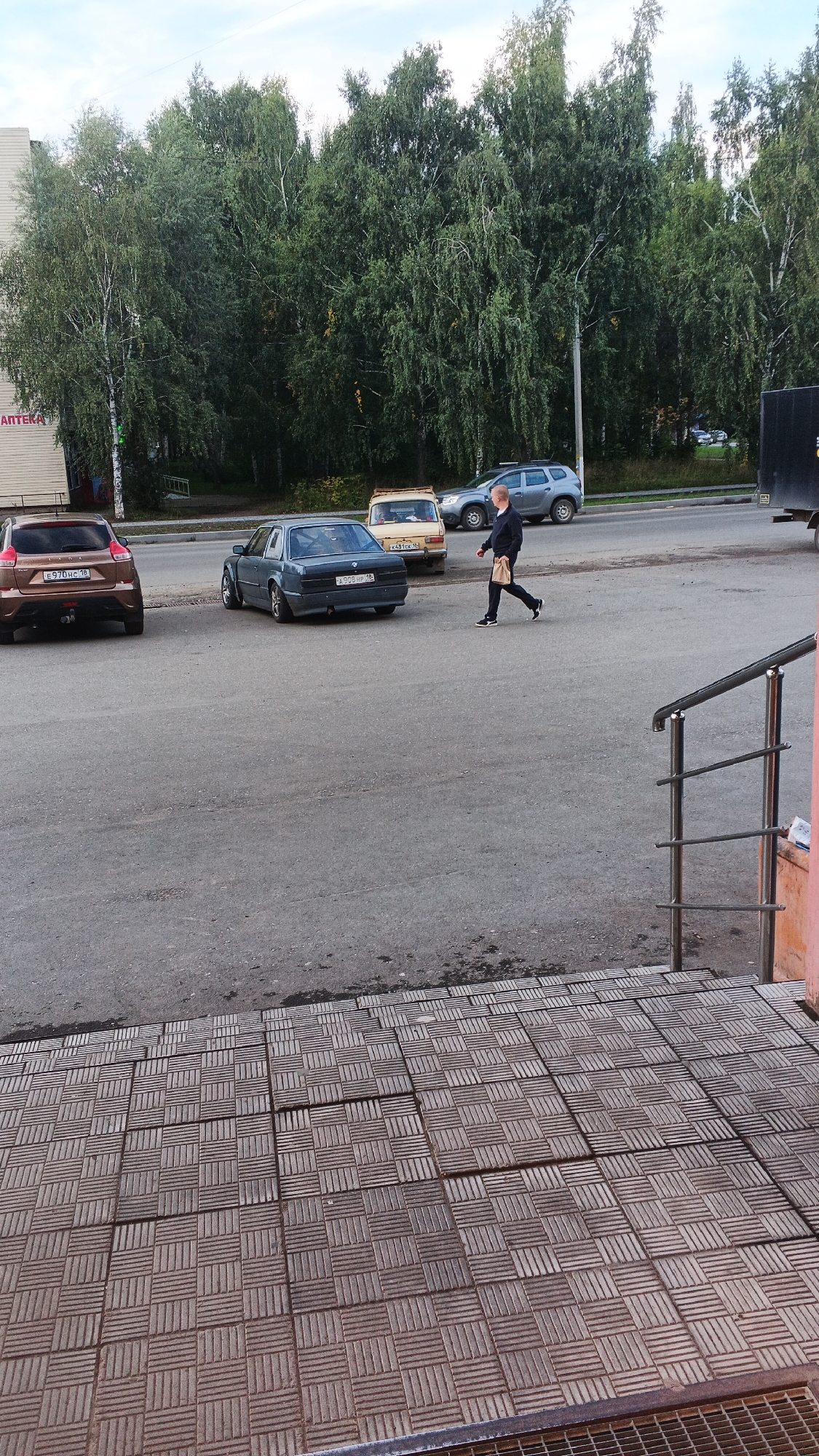 A man walks across a paved area in front of a building. He is carrying a brown paper bag and is wearing a black jacket, black pants, and white-soled shoes. Several cars are parked on the street behind him, including a dark blue sedan, a tan sedan, and a silver SUV. Tall trees line the street. A metal railing is visible in the foreground. The scene appears to be in an urban or suburban area of Izhevsk, Russia.