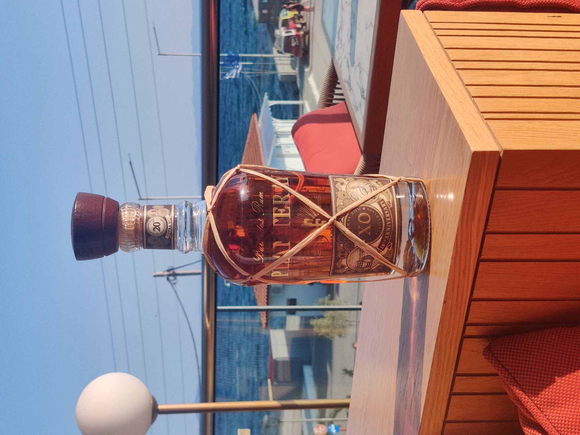 A bottle of Mount Gay XO 20th Anniversary rum sits on a wooden table on a balcony overlooking the sea in Sithonia, Greece. The rum bottle is made of glass and is filled with amber-colored liquid. It is tied with a straw net. The label on the bottle reads "Mount Gay Rum" and "XO 20th Anniversary". In the background, the sea is visible, with boats and buildings on the shore. A white spherical lamp is to the left of the bottle. The sky is blue and clear. The overall scene is one of leisure and relaxation.
