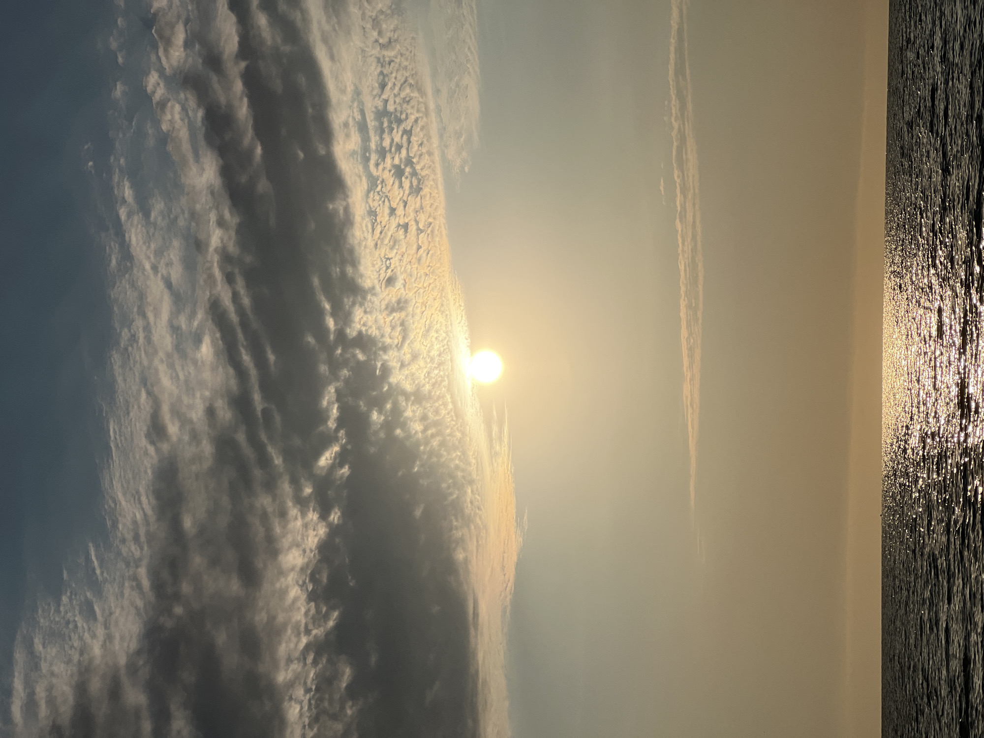 The image depicts a serene seascape under a partly cloudy sky. The sun is visible, partially obscured by clouds, casting a warm, golden light across the water. The sea is calm, with gentle waves reflecting the sunlight. In the distance, a faint silhouette of a boat can be seen on the horizon. The sky is a gradient of blues and grays, with a mix of dark and light clouds creating a textured pattern. The overall atmosphere is peaceful and tranquil. The location in Borovoe, Russia, is not discernible from the image itself, as it is a general seascape.