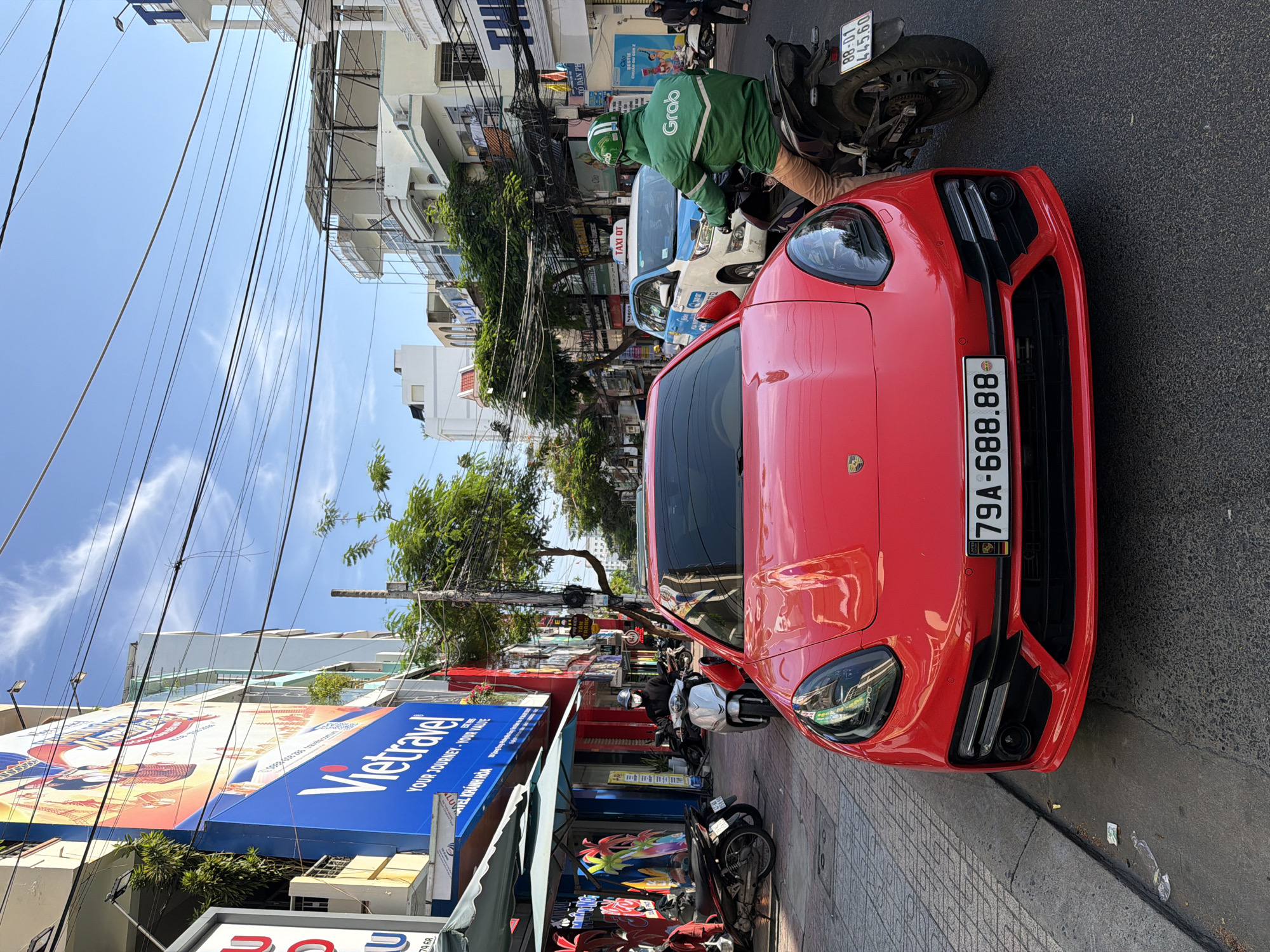 In Nha Trang, Vietnam, a vibrant street scene unfolds under a clear blue sky. A striking red Porsche Panamera is parked on the right side of the road, its front bumper facing the camera. The car's license plate reads "79A-688.88".
To the right of the Porsche, a Grab delivery driver, clad in a distinctive green jacket and helmet, is seated on a black motorcycle. The motorcycle's license plate is "88-D1 445.60".
In the background, a white and blue taxi with "TAXI OT" visible on its roof is also present. The street is lined with buildings, some adorned with colorful advertisements, including a large "Vietravel" banner. Trees and power lines crisscross the sky, adding to the urban ambiance. Several other motorcycles are parked along the sidewalk. A person wearing a mask and helmet is on a silver scooter to the left of the Porsche. The scene captures a typical day in this bustling Vietnamese city.