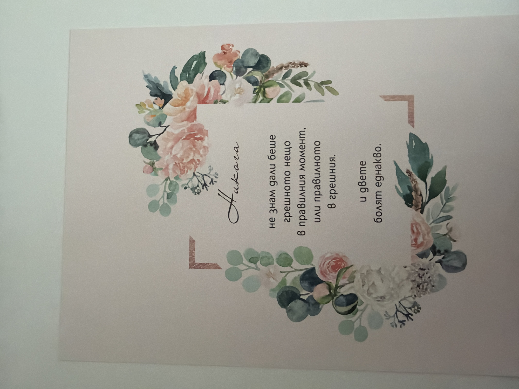The image displays a card with floral watercolor illustrations. The text on the card is in Bulgarian. At the top center, the word "Никога" (Never) is written in a stylized font. Below it, a quote reads: "не знам дали беше грешно нещо в правилния момент, или правилното в грешния. И двете болят еднакво." (I don't know if it was something wrong at the right moment, or the right thing at the wrong one. Both hurt equally.) The card is set against a pale, peachy-pink background and framed by a delicate watercolor wreath of pink and white roses, eucalyptus leaves, and other greenery. The lighting suggests it's indoors, likely on a flat surface. There are no people or signs of the city of Pazardzhik, Bulgaria, present in the image.