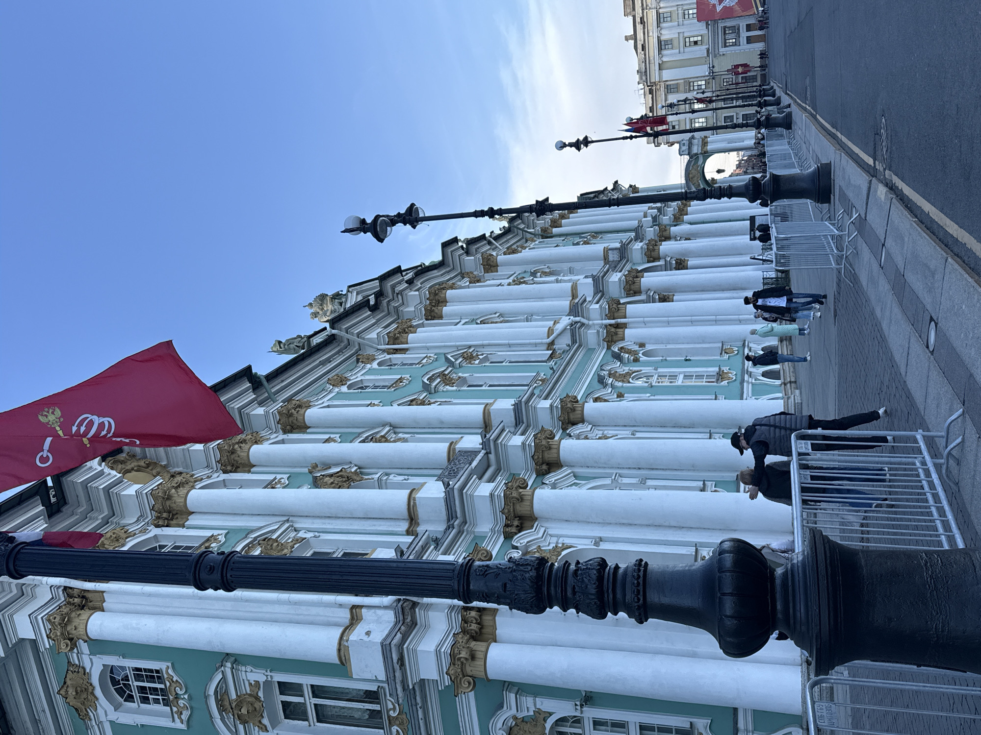 The image captures a vibrant street scene dominated by a grand, ornate historical building, characteristic of Saint Petersburg, Russia. The building, likely the Winter Palace, features a distinctive light turquoise facade with numerous white columns, adorned with elaborate gold Corinthian capitals and bases, and other decorative elements. Sculptural details are visible along the roofline.
A large red flag with the golden two-headed eagle emblem of Russia (including a scepter and orb) flies prominently from the building's upper left section.
Along the right side of the building, a wide paved sidewalk is present, lined with ornate black streetlights. White metal crowd control barriers run along the edge of the sidewalk, separating pedestrians from the asphalt road beyond. Several people are visible on the sidewalk, walking or standing, some near the barriers. In the distance, other historical buildings and an arched structure can be seen.
The sky is a clear, bright blue, indicating good weather conditions during daytime. Visible Cyrillic text on one of the white barriers, "СПб ГБУ," identifies it as belonging to a Saint Petersburg State Budgetary Institution, confirming the location. The scene portrays a bustling urban environment amidst impressive historical architecture.