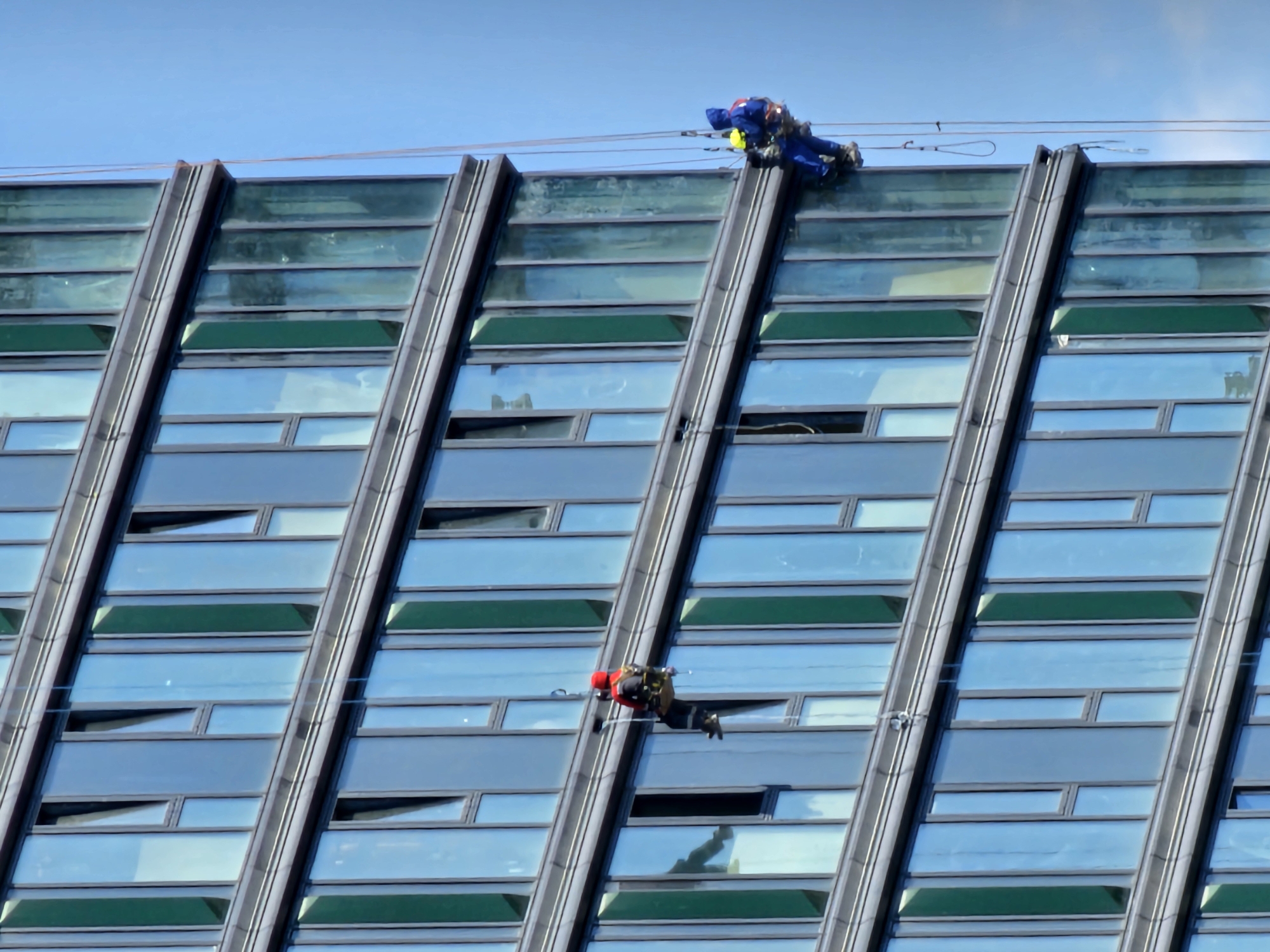 The image shows two window cleaners on the side of a modern glass building. The building has a facade of rectangular glass panels, some of which are slightly ajar. The sky is blue and clear. One cleaner, wearing a red hard hat and a grey and red uniform, is suspended by ropes on the left side of the building, appearing to be working on a window. Further to the right, another cleaner, wearing a blue uniform and a yellow hard hat, is also suspended by ropes, positioned higher on the building and seemingly working on a different section of windows. The scene is a typical urban environment, suggesting commercial or residential high-rise construction or maintenance. No specific identifying features place this scene in Moscow, Russia.