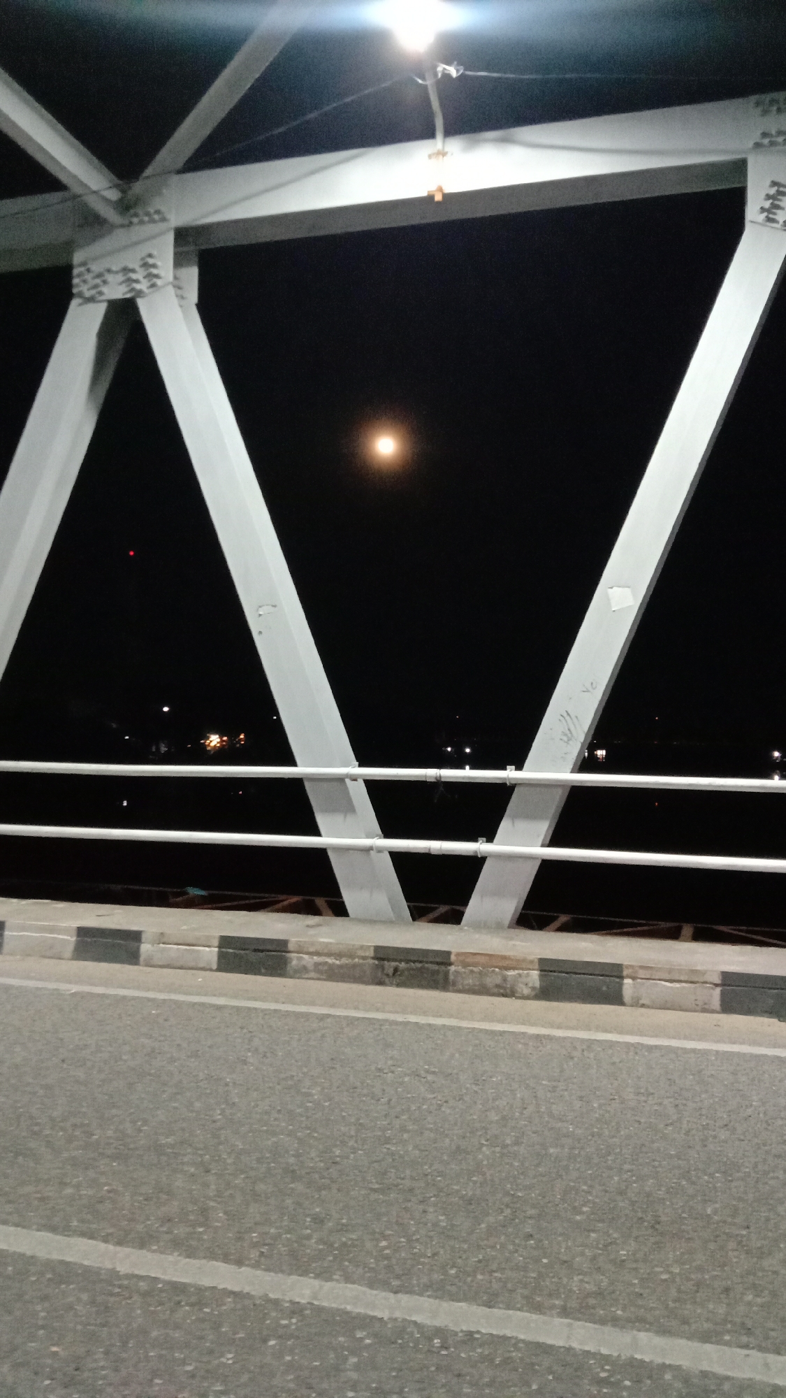 The image depicts a night scene on a bridge in Lhokseumawe, Indonesia. The photograph is taken from street level, showing the asphalt road with white lane markings and a sidewalk curb painted with alternating black and gray blocks. A safety railing composed of two white horizontal bars is visible. The bridge's metal structure, composed of white triangular supports, dominates the upper part of the frame. A bright light illuminates the top of the structure. In the center, a light source, possibly the moon or another light source, appears against the dark night sky. Several faint lights from the cityscape are present on the horizon.