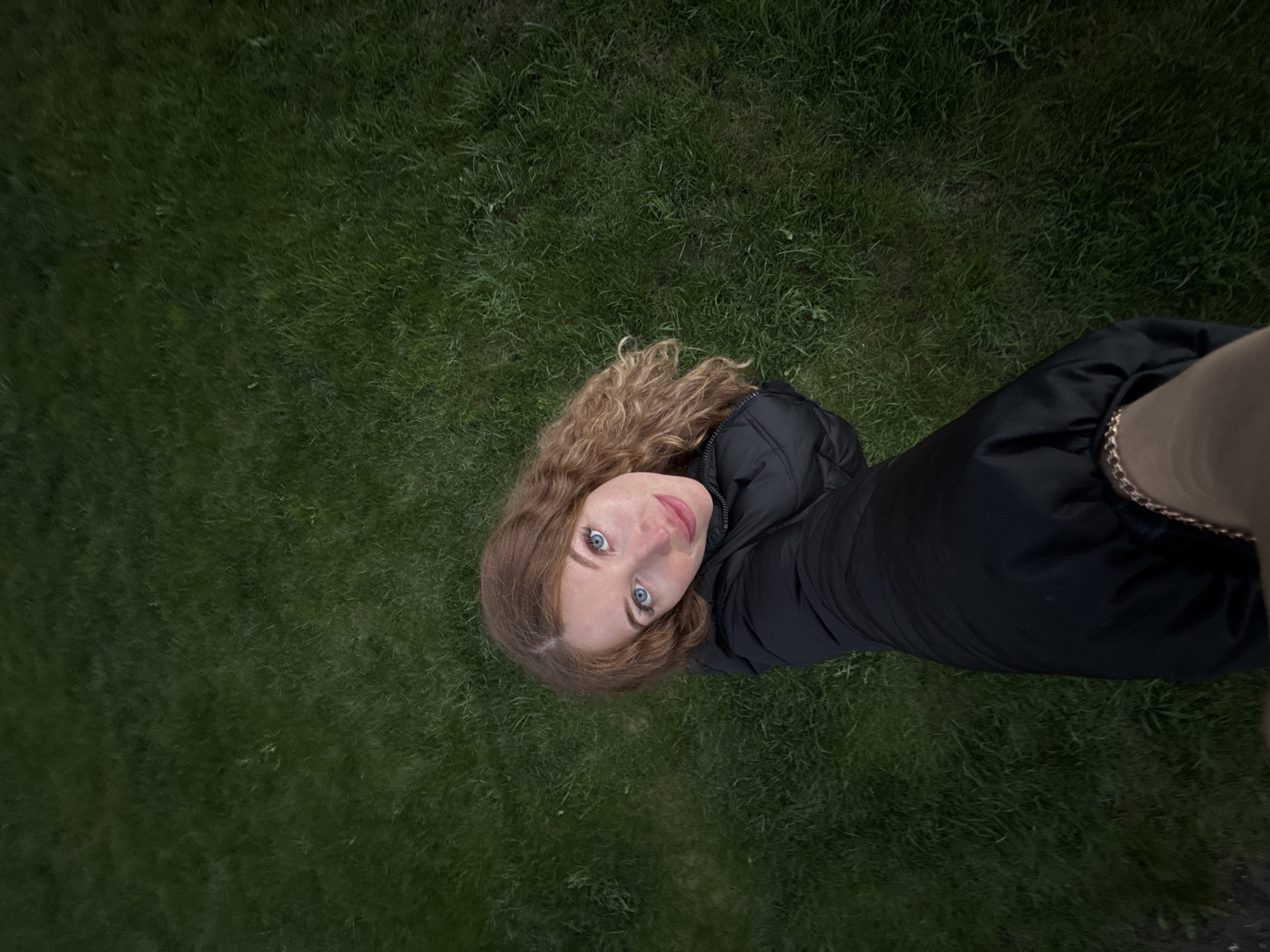 A young woman with light brown, wavy hair and blue eyes is lying on a patch of green grass. She is wearing a black jacket. Her head is tilted upward, looking toward the camera. A small part of what appears to be a beige-colored sleeve with a gold chain detail is visible on the right side of the frame. The photograph appears to be taken from directly above her, giving a skewed perspective. The setting appears to be outdoors on a lawn or grassy area. The location in the city of Chaikovsky, Russia, cannot be confirmed from the provided image alone.