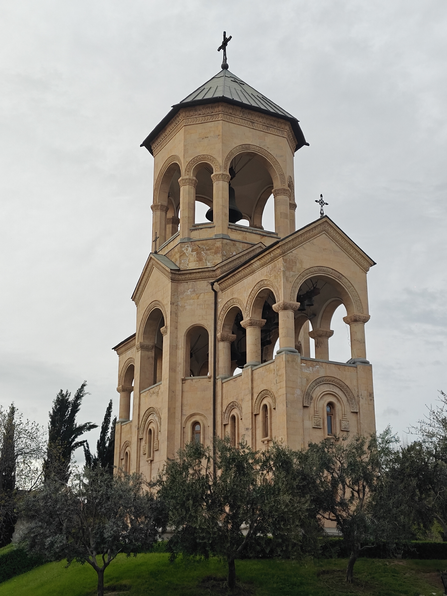 The image displays a majestic bell tower or church facade, indicative of Georgian Orthodox architecture, set against a bright, overcast sky. The structure is built from light-colored, possibly sandstone, and exhibits multiple levels of ornate arched openings supported by columns.
The uppermost section is an octagonal bell tower featuring a dark, conical roof crowned by a prominent black Orthodox cross. Large bells are visible hanging within the arches of this tier. Below this, a section with a gable roof also showcases arched openings, revealing more bells inside. A smaller black Orthodox cross adorns the peak of this gable. The lower parts of the building continue the architectural style with arched windows and elaborate stone carvings, including decorative patterns above the arches.
The foreground features a lush, green lawn sloping gently upwards, dotted with various trees. Several mature, dark-leaved trees, possibly olive trees, occupy the lower half of the frame, partially obscuring the base of the structure. Taller, slender evergreen trees are visible towards the left side of the frame.
The image captures the building under the diffuse light of a cloudy day. No people or specific activities are present. The architectural style strongly suggests its location in Tbilisi, Georgia, likely the bell tower of the Holy Trinity Cathedral of Tbilisi (Sameba Cathedral). No visible text is present in the image.