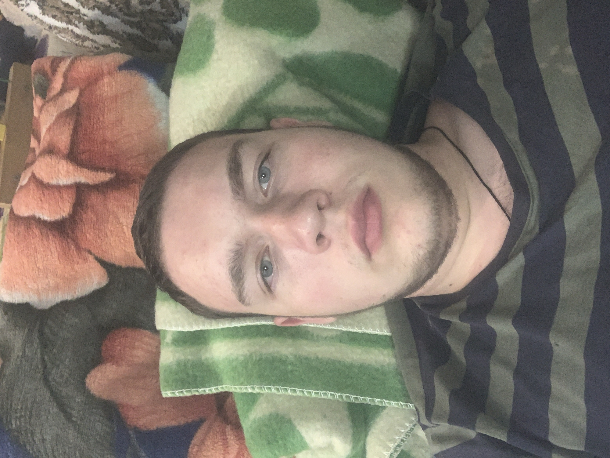 A young adult male is lying down, facing the camera. He has short brown hair, blue eyes, and a light beard. He is wearing a dark blue and green striped t-shirt and a thin black cord necklace. He is lying on a green and white striped blanket, which is on top of other blankets. The background blankets are dark grey and have an orange, floral pattern. The scene appears to be indoors, possibly in a bedroom. The image does not provide any specific clues about the location within Panteleevka, Russia.